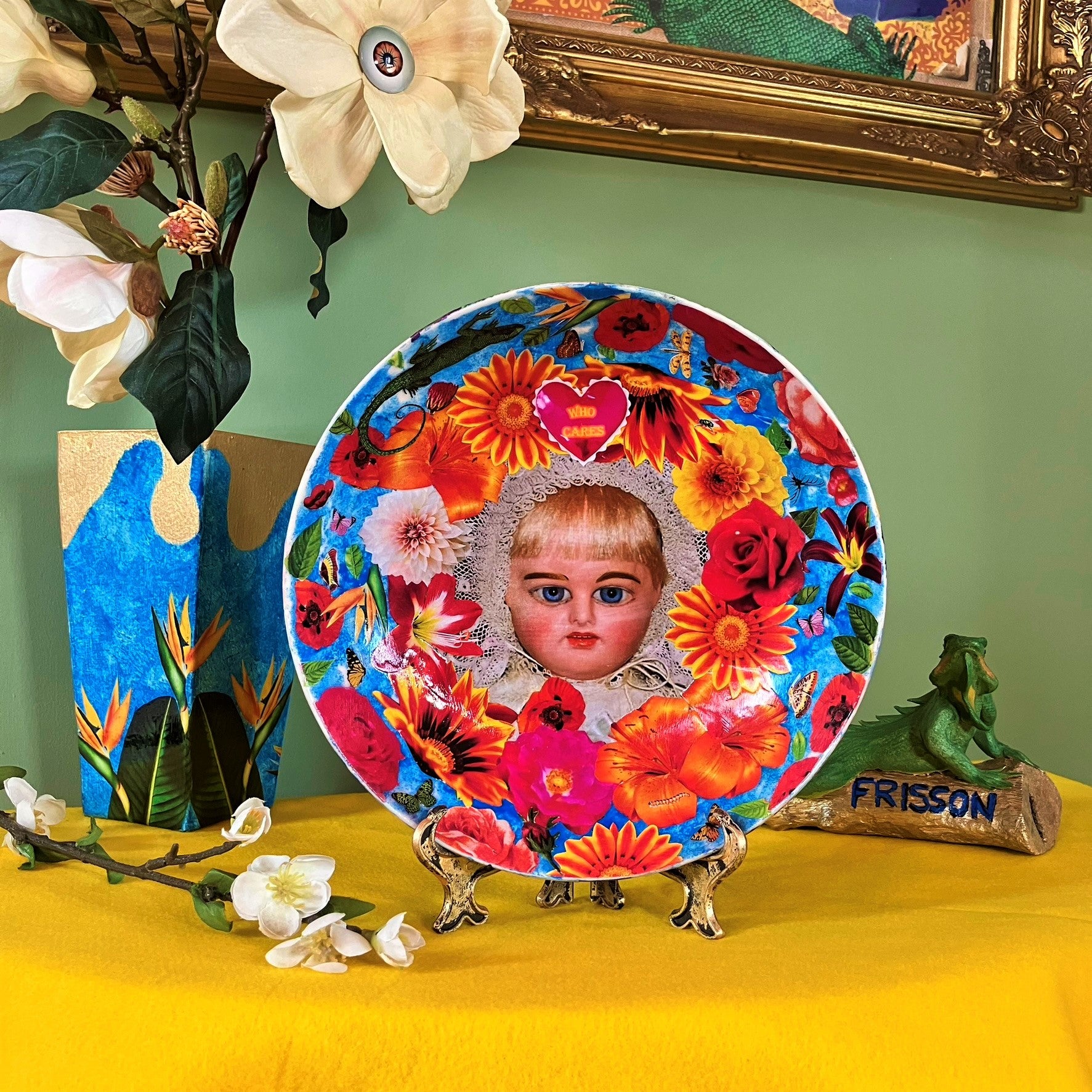 "Who Cares" Wall Plate by House of Frisson, featuring a vintage doll surrounded by flowers, moths, and a lizard, on a blue background. Plate on a plate stand, resting on a table.