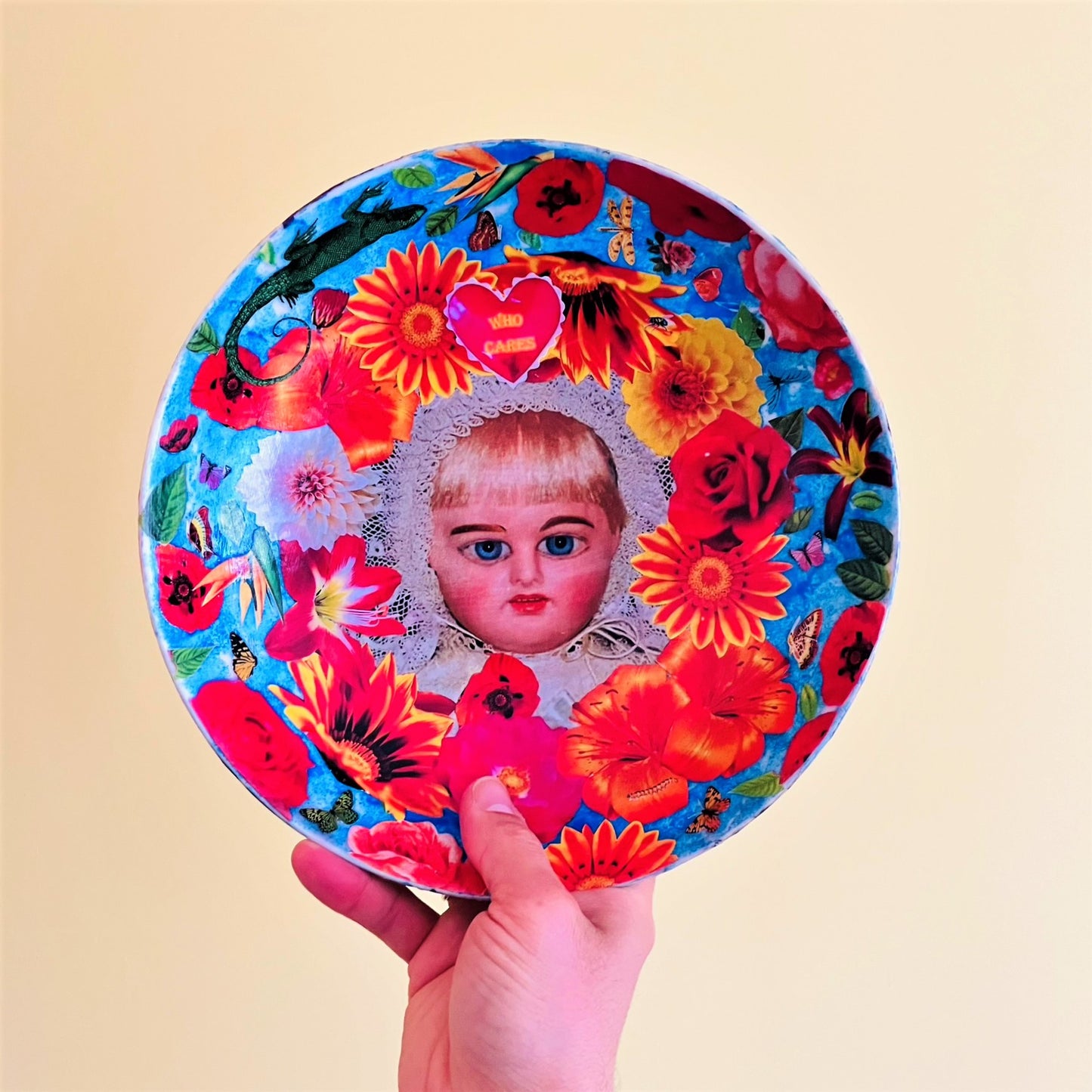 "Who Cares" Wall Plate by House of Frisson, featuring a vintage doll surrounded by flowers, moths, and a lizard, on a blue background. Holding the plate.