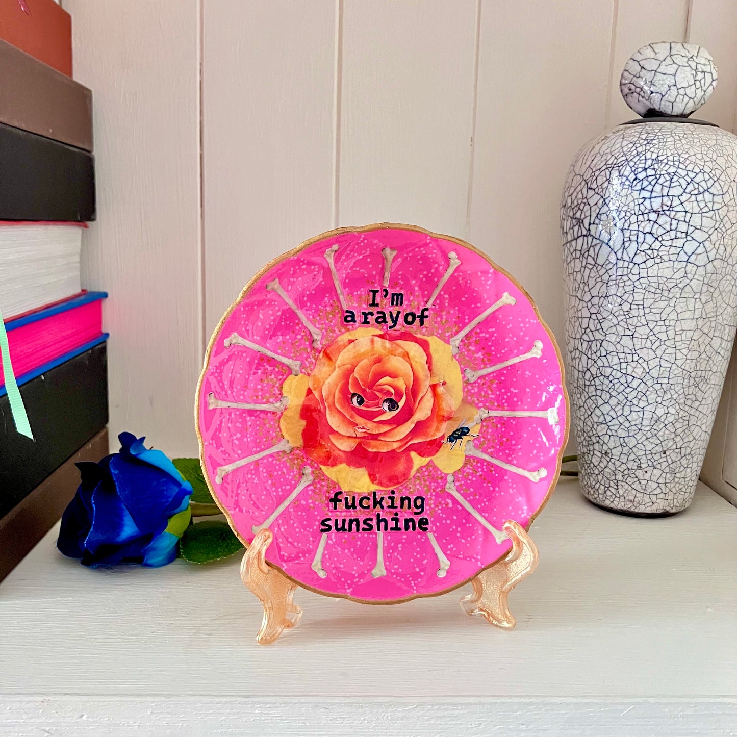 "I'm a Ray of Fucking Sunshine" Pink Wall Plate by House of Frisson, featuring an orange rose with eyes, bones, and a fly. Plate on a plate stand resting on a shelf.