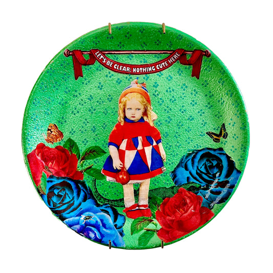 "Let's Be Clear: Nothing Cute Here" Wall Plate by House of Frisson, featuring a collage artwork of a vintage doll surrounded by roses, moths, and a lizard, on a metallic green background.