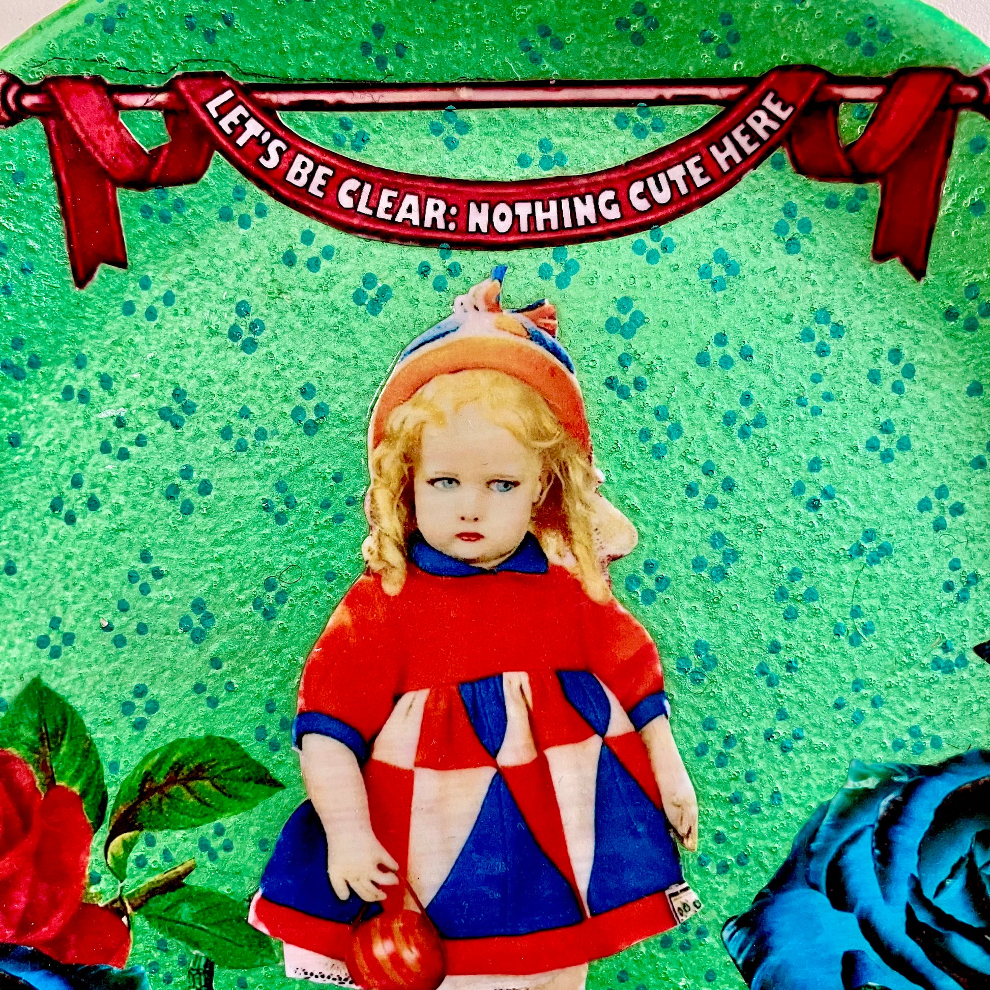 "Let's Be Clear: Nothing Cute Here" Wall Plate by House of Frisson, closeup detail of the collage, showing a vintage doll, and roses, on a metallic green background.