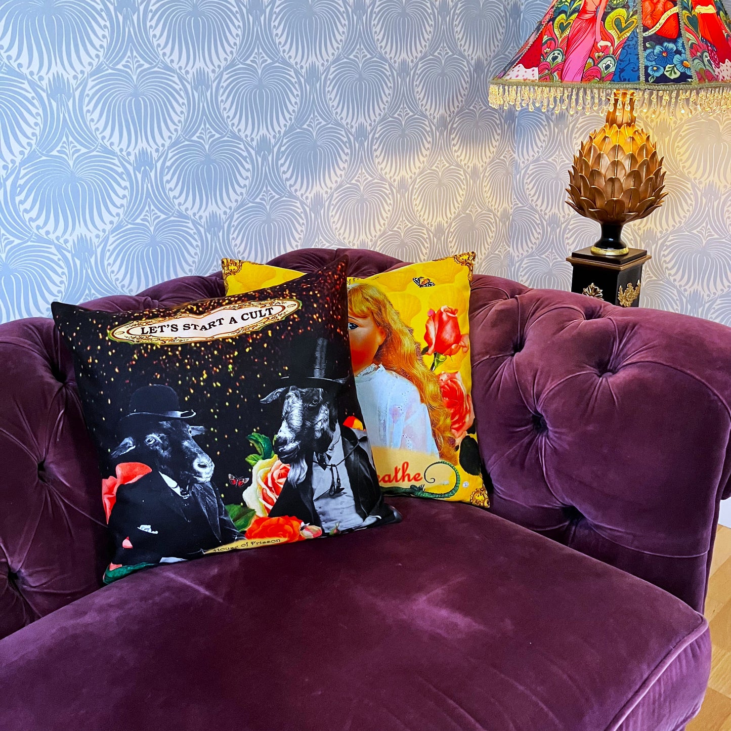 "Let's Start A Cult" Cushion Cover by House of Frisson, featuring a collage of two Victorian-dressed  glentlemen with goat heads, surrounded by colourful flowers, against a black background. Cushion on a sofa with other cushions and ornaments.