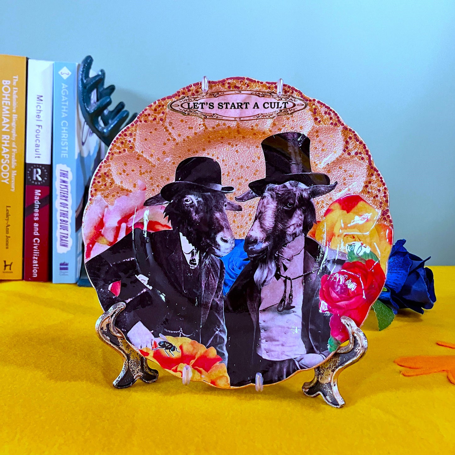 Gold upcycled wall plate by House of Frisson, featuring two Victorian gentlemen with goat heads surrounded by colourful flowers and a fly. Plate on a plate stand resting on a table.