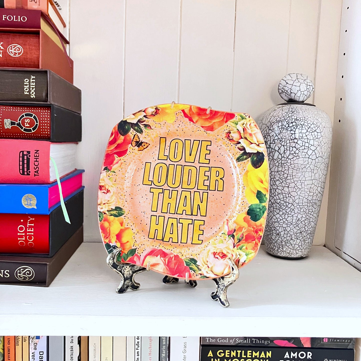 "Love Louder Than Hate" Upcycled Wall Plate by House of Frisson, featuring a collage of the words "love louder than hate" set against a peach background and framed by roses. Plate on a plate stand resting on a shelf.