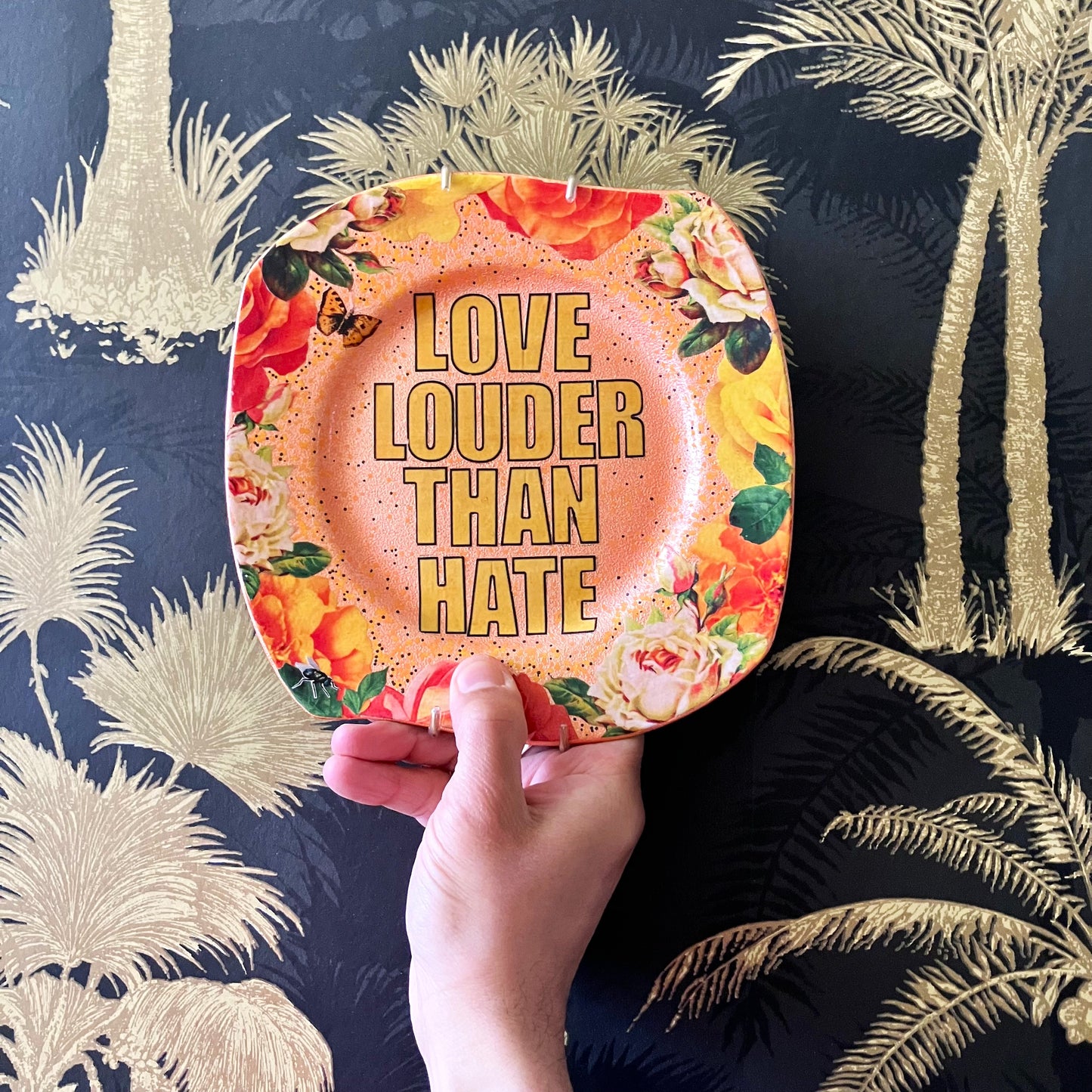 "Love Louder Than Hate" Upcycled Wall Plate by House of Frisson, featuring a collage of the words "love louder than hate" set against a peach background and framed by roses. Showing a closeup of someone holding the plate.