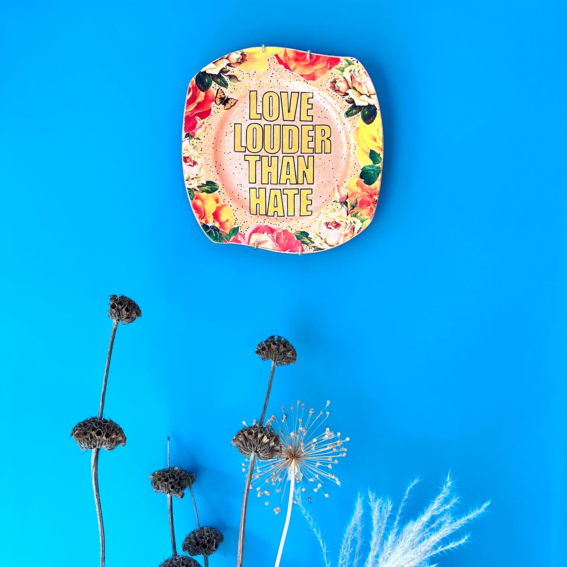 "Love Louder Than Hate" Upcycled Wall Plate by House of Frisson, featuring a collage of the words "love louder than hate" set against a peach background and framed by roses. Plate hanging on a blue wall.
