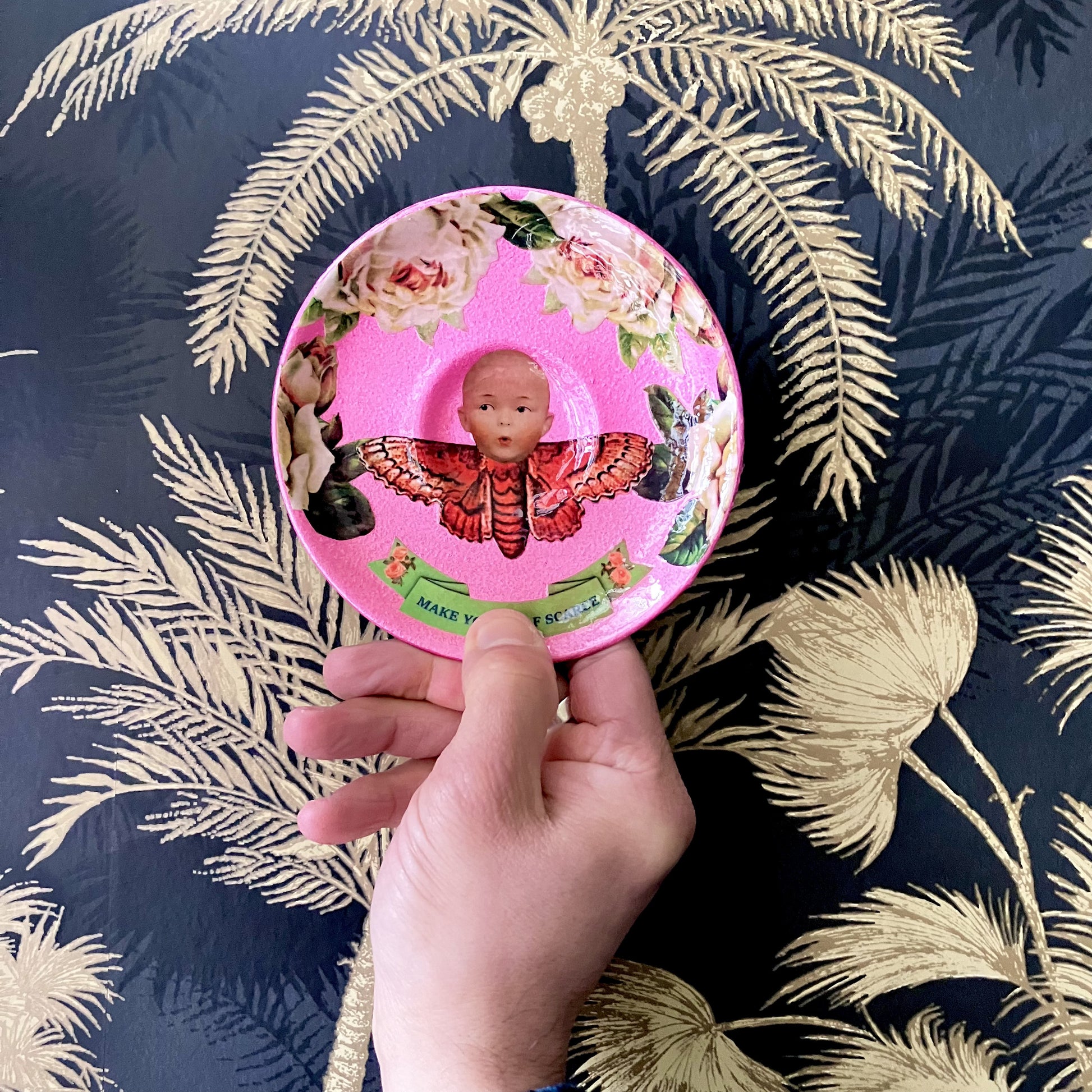 "Make Yourself Scarce" Trinket Dish by House of Frisson. Featuring a moth with doll head, framed by roses, on a pink background. Holding dish against a wall.