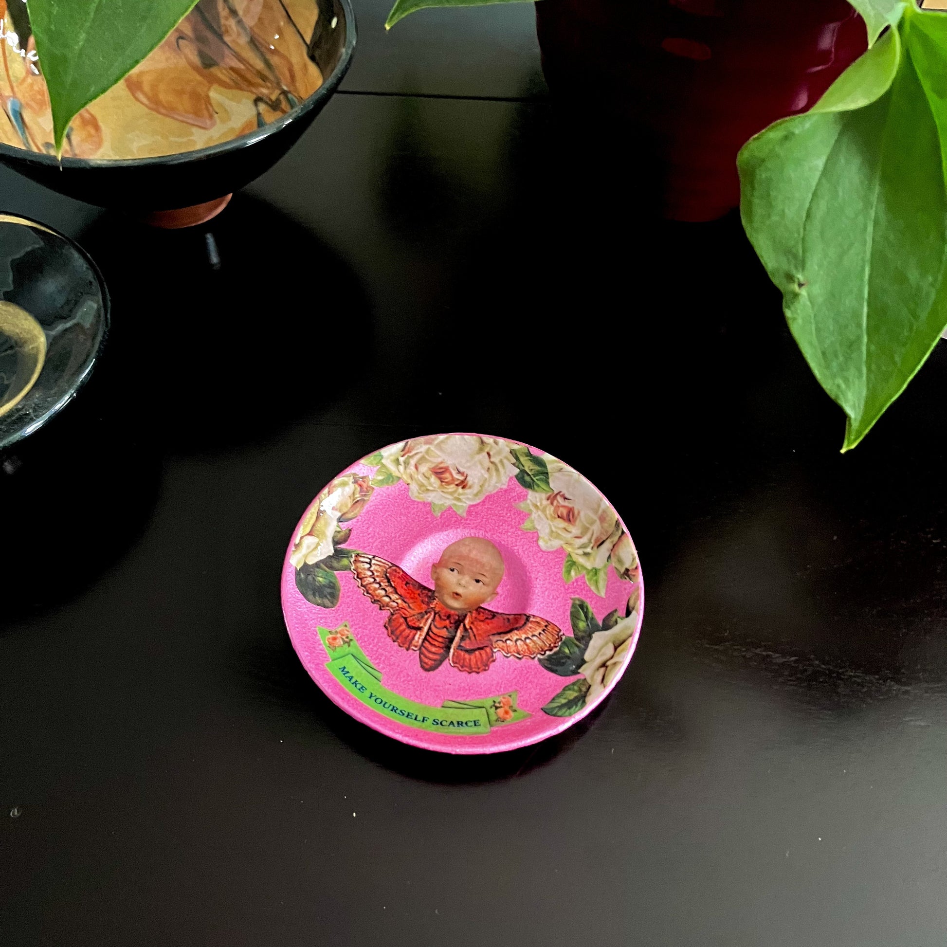 "Make Yourself Scarce" Trinket Dish by House of Frisson. Featuring a moth with doll head, framed by roses, on a pink background. Dish on a coffee table.