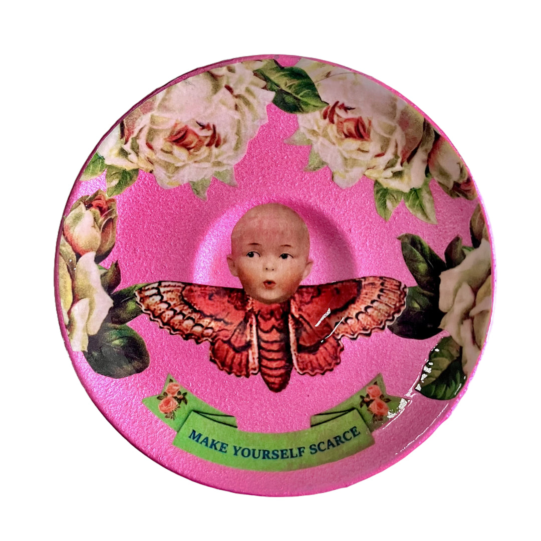"Make Yourself Scarce" Trinket Dish by House of Frisson. Featuring a moth with doll head, framed by roses, on a pink background.