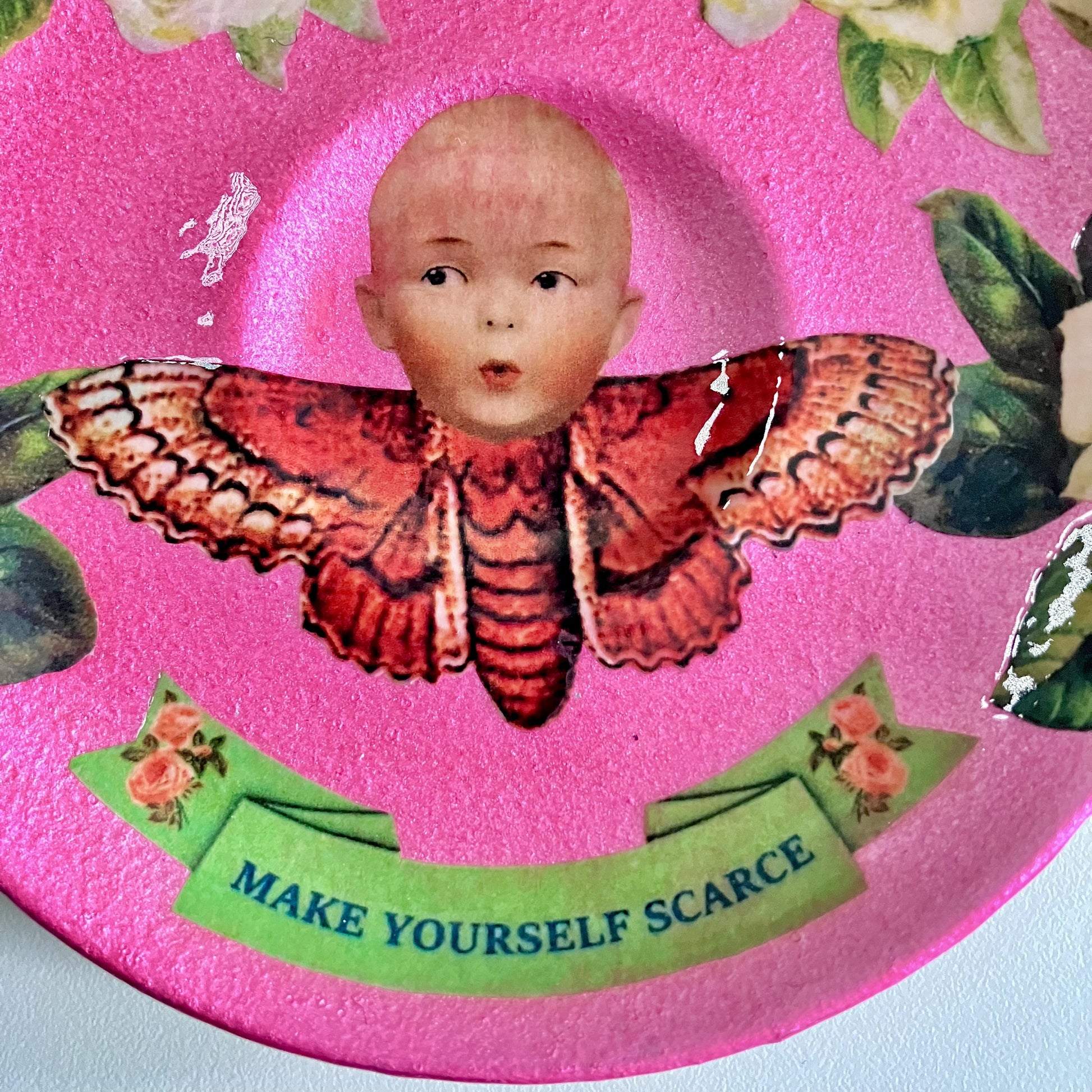 "Make Yourself Scarce" Trinket Dish by House of Frisson. Featuring a moth with doll head, framed by roses, on a pink background. Closeup detail showing a moth with doll head.