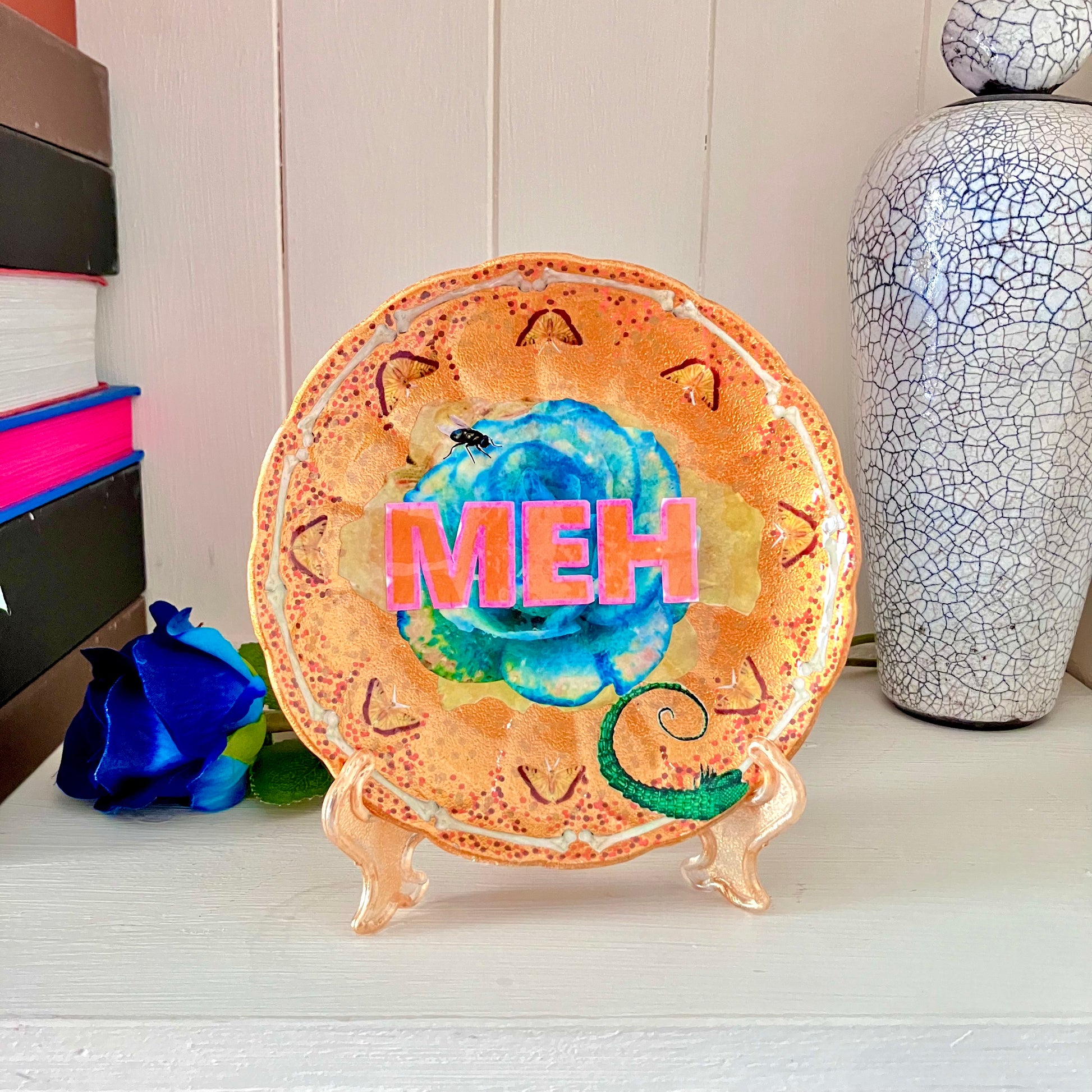 "Meh" upcycled Wall Plate by House of Frisson, featuring a green rose, some moths, bones, and House of Frisson's creatures. Plate on a plate stand resting on a shelf.