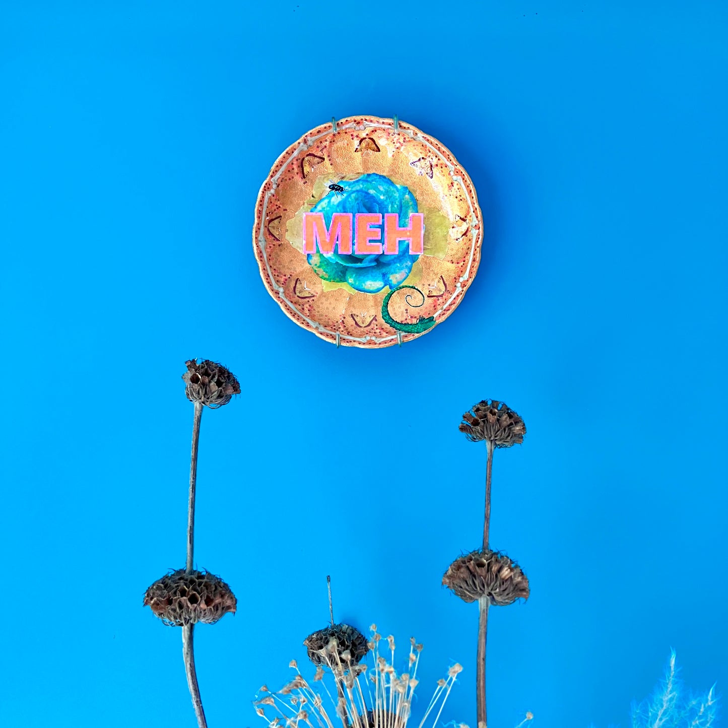 "Meh" upcycled Wall Plate by House of Frisson, featuring a green rose, some moths, bones, and House of Frisson's creatures. Showing plate hanging on a blue wall.