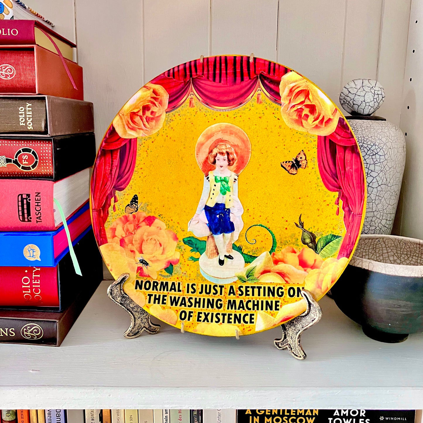 "Normal Is Just A Setting On The Washing Machine Of Existence" Wall Plate by House of Frisson, on a plate stand resting on a bookshelf.