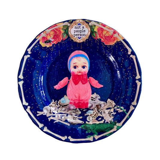 "Not A People Person" dark blue wall plate by House of Frisson, featuring a vintage celluloid doll surrounded by human bones and pearls.