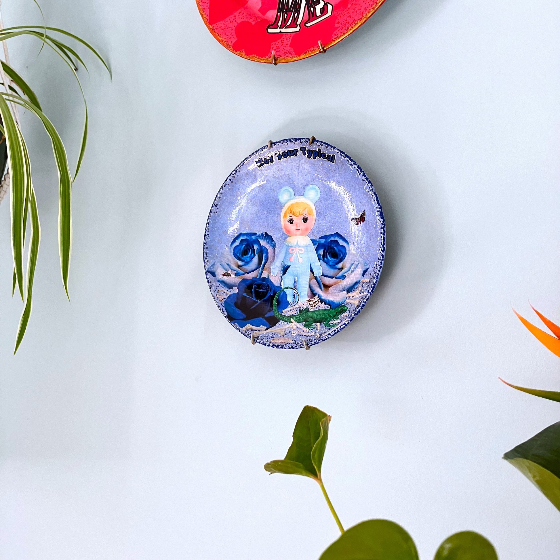 Light Blue Wall Plate by House of Frisson, featuring a kitschy vintage rubber doll surrounded by blue roses, with scattered bones, a lizard, and a fly. Guaranteed to bring a dose of fun and personality to your space!  Plate hanging on a wall.