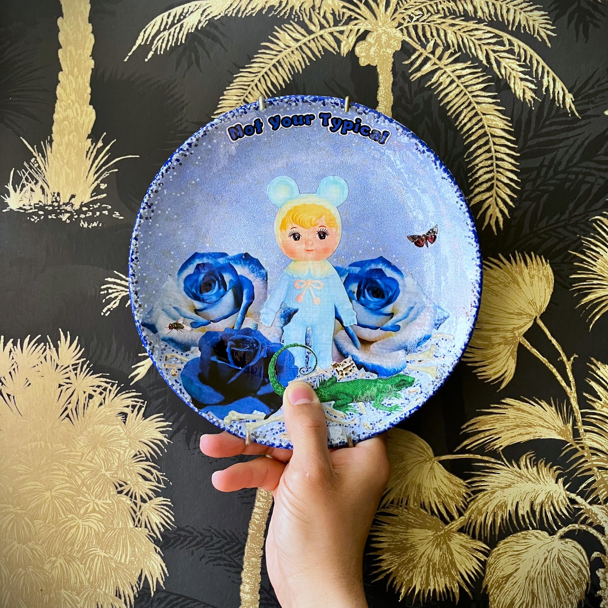 Light Blue Wall Plate by House of Frisson, featuring a kitschy vintage rubber doll surrounded by blue roses, with scattered bones, a lizard, and a fly. Guaranteed to bring a dose of fun and personality to your space!  Holding the plate against a wall.