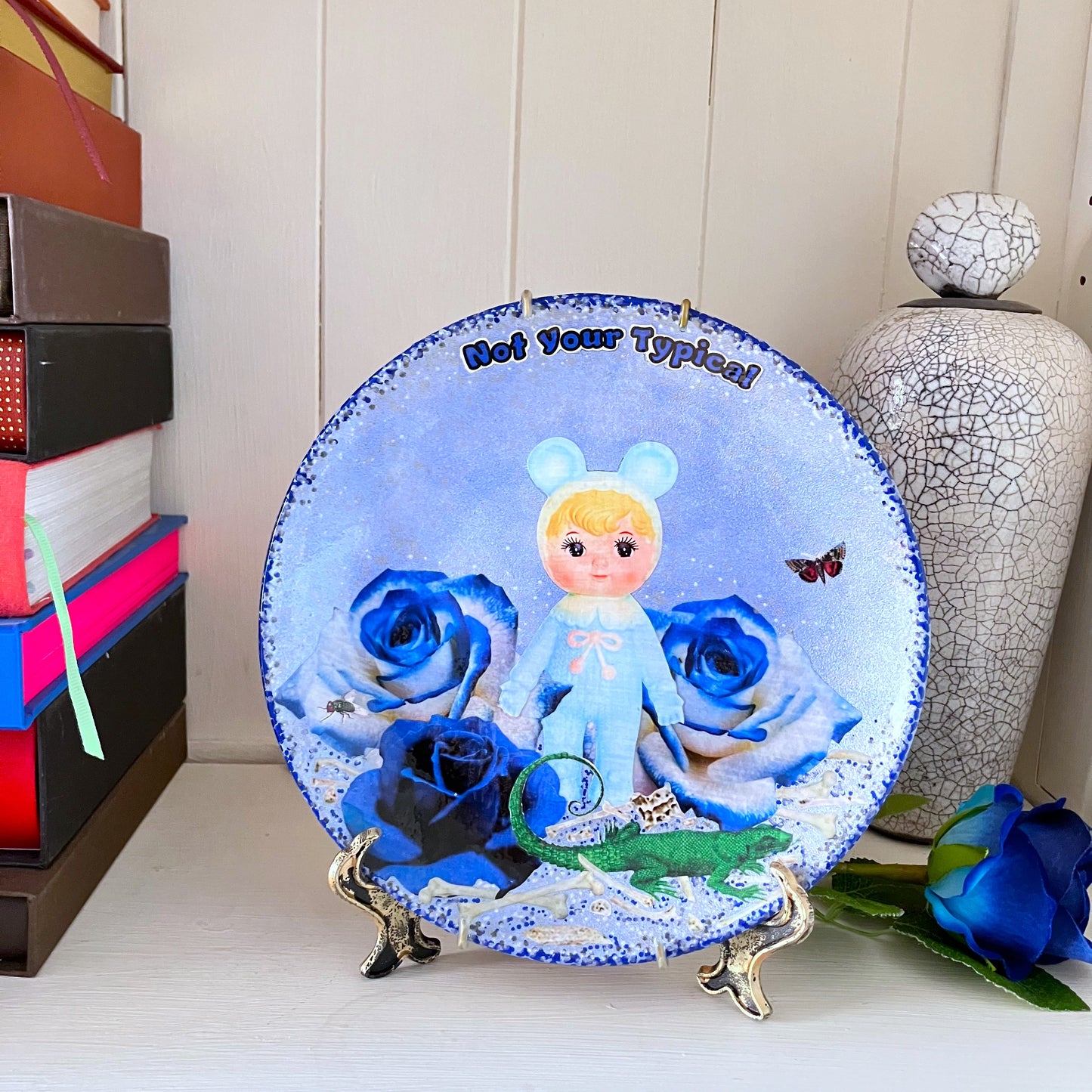 Light Blue Wall Plate by House of Frisson, featuring a kitschy vintage rubber doll surrounded by blue roses, with scattered bones, a lizard, and a fly. Guaranteed to bring a dose of fun and personality to your space!  Plate on a plate stand resting on a shelf.