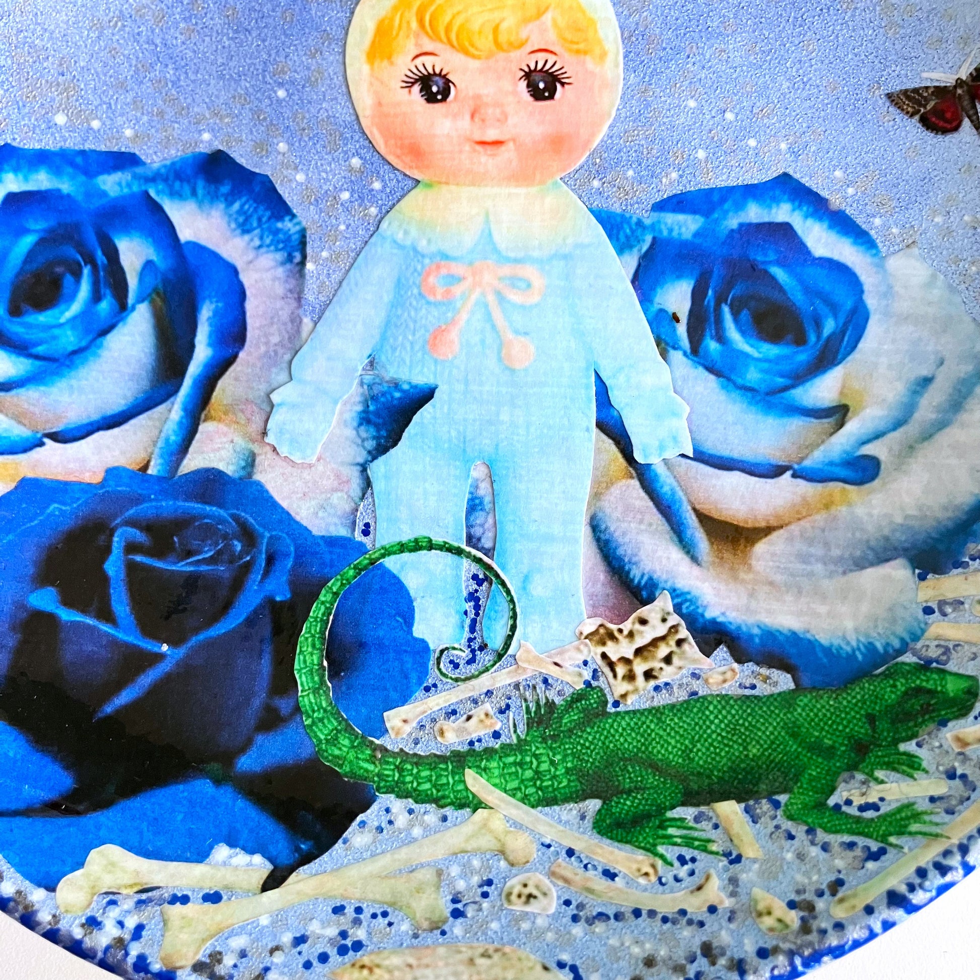 Light Blue Wall Plate by House of Frisson, featuring a kitschy vintage rubber doll surrounded by blue roses, with scattered bones, a lizard, and a fly. Guaranteed to bring a dose of fun and personality to your space!  Closeup detail showing the collage.