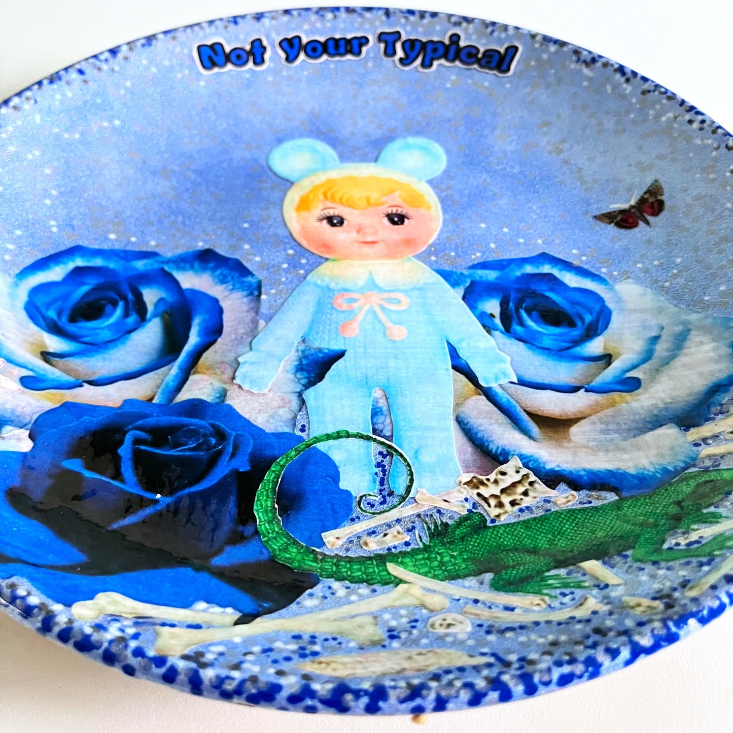 Light Blue Wall Plate by House of Frisson, featuring a kitschy vintage rubber doll surrounded by blue roses, with scattered bones, a lizard, and a fly. Guaranteed to bring a dose of fun and personality to your space!  Closeup detail showing the collage.