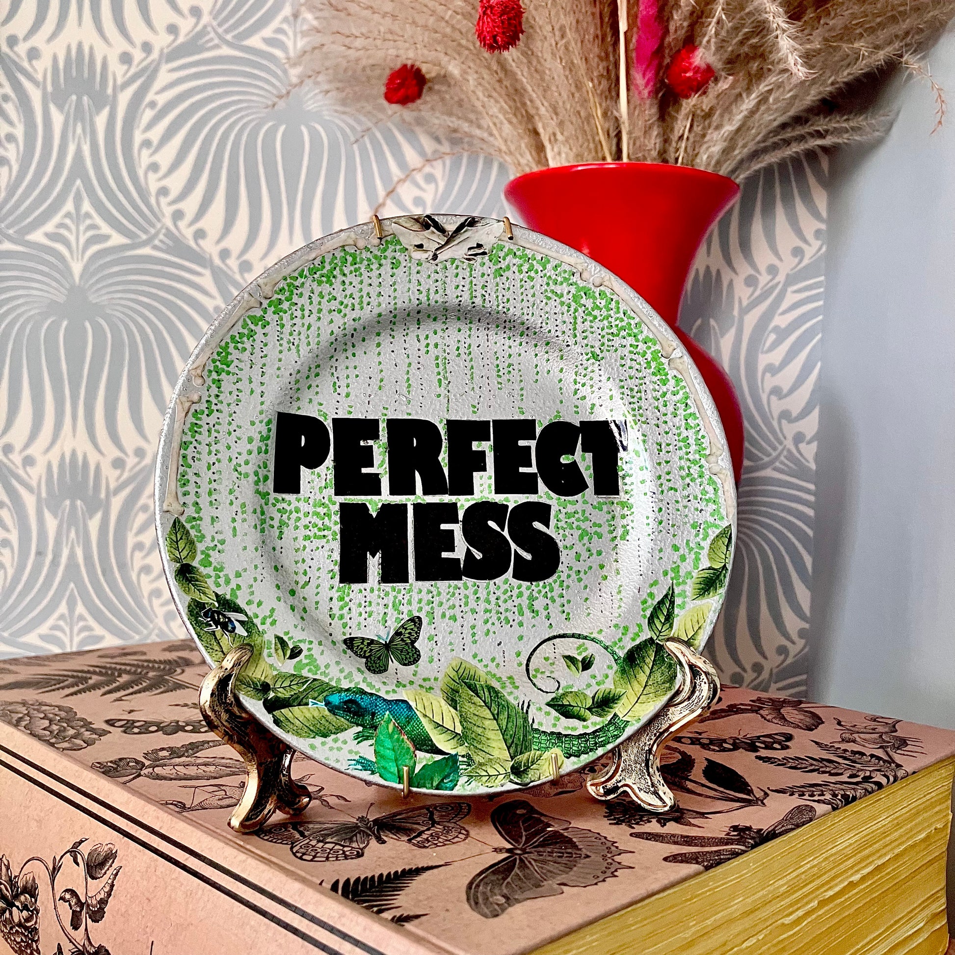 "Perfect Mess" Wall Plate by House of Frisson, on a plate stand, resting on a bookshelf.