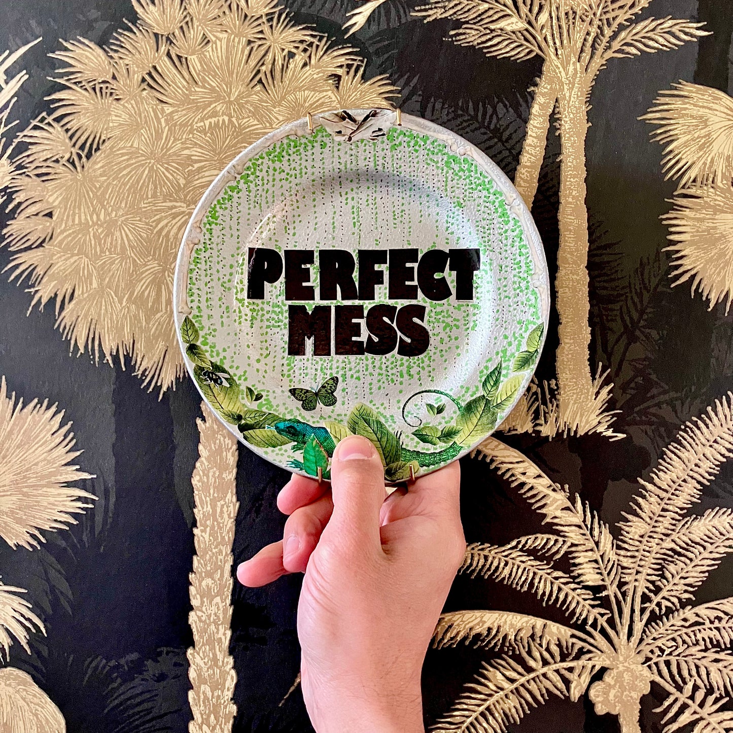 "Perfect Mess" Wall Plate by House of Frisson, on hand, against a wallpaper.
