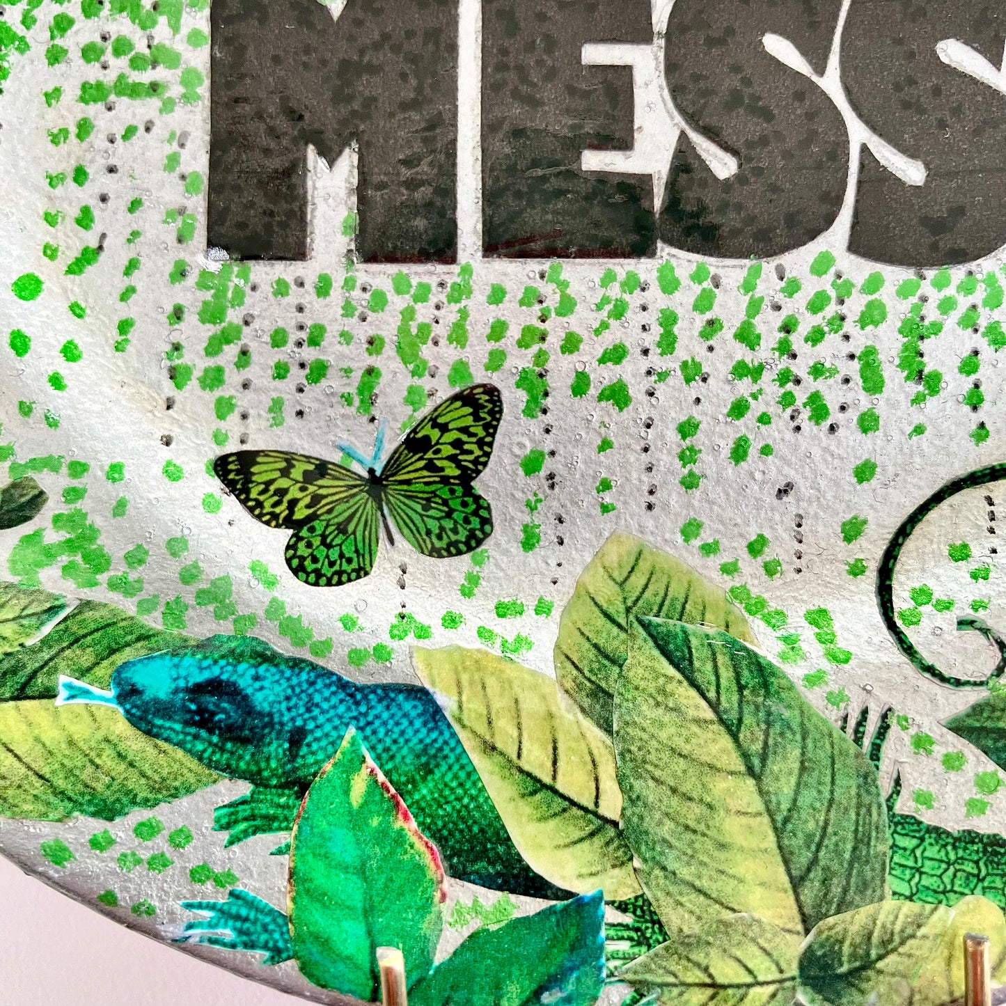 "Perfect Mess" Wall Plate by House of Frisson, closeup detail of the collage showing green leaves, a lizard, a moth, and green pattern on a silver background.