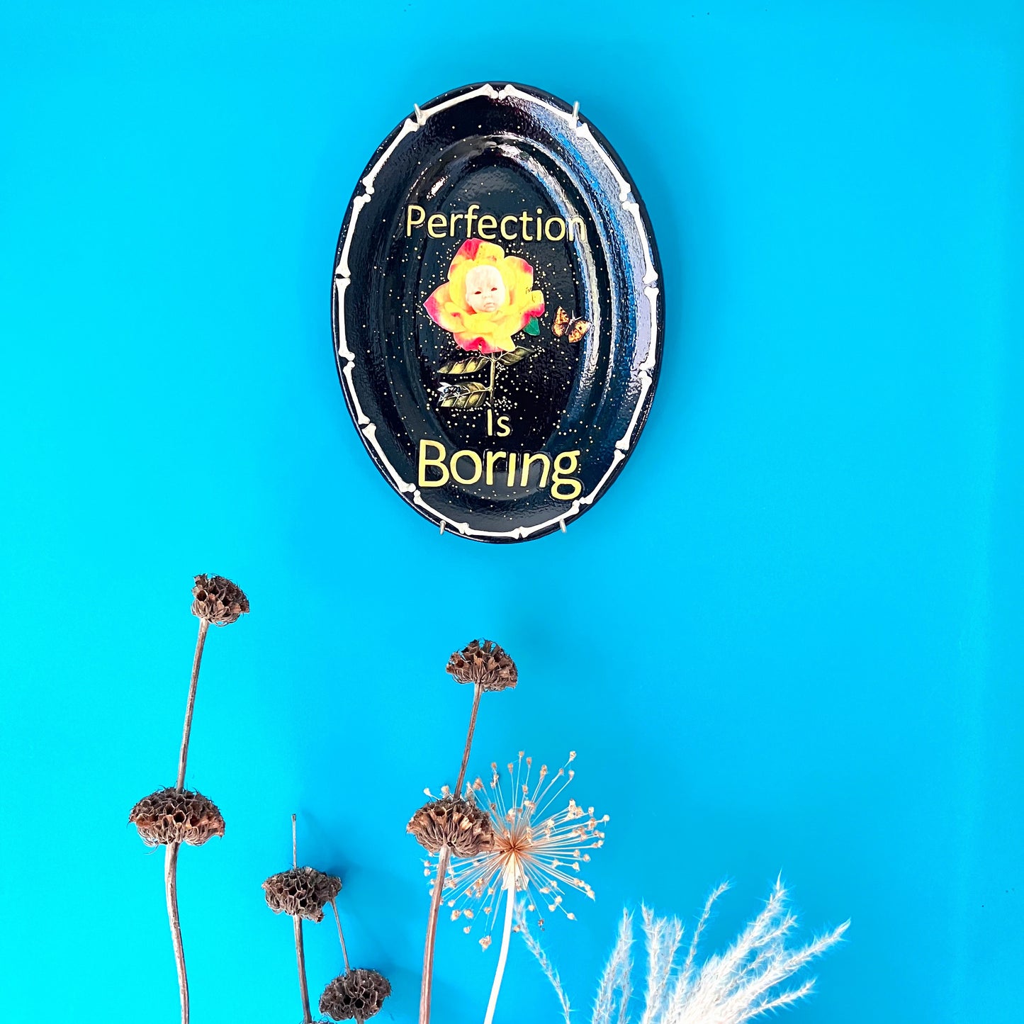 "Perfection Is Boring" Upcycled Wall Plate by House of Frisson, featuring a collage of a yellow rose with a doll face, a moth, and House of Frisson's fly, in a frame of bones. Plate hanging on a blue wall.