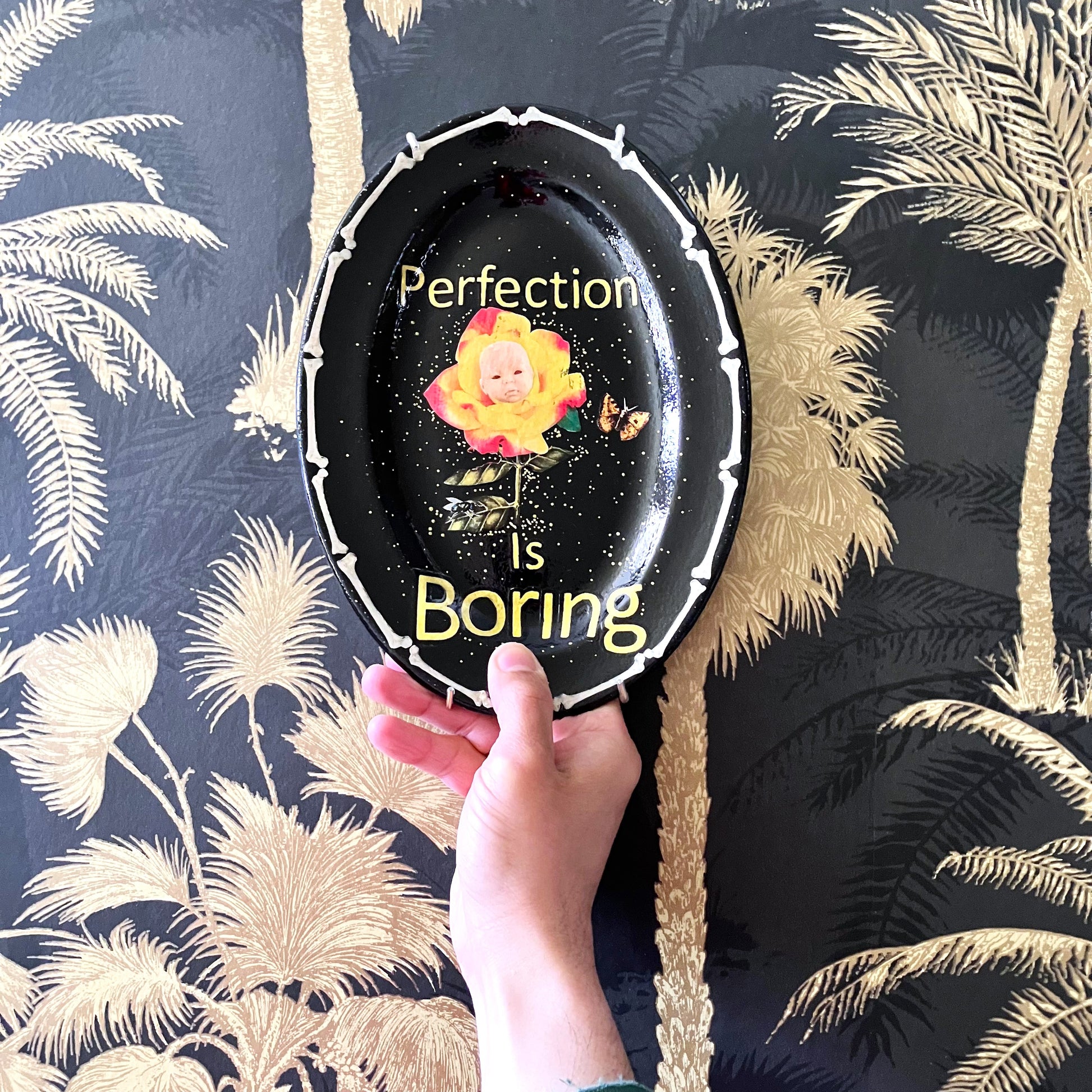 "Perfection Is Boring" Upcycled Wall Plate by House of Frisson, featuring a collage of a yellow rose with a doll face, a moth, and House of Frisson's fly, in a frame of bones. Closeup image showing someone holding the plate.