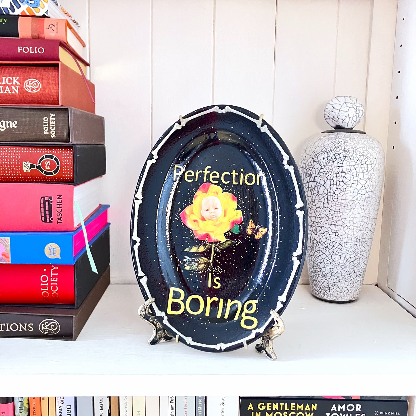 "Perfection Is Boring" Upcycled Wall Plate by House of Frisson, featuring a collage of a yellow rose with a doll face, a moth, and House of Frisson's fly, in a frame of bones. Plate on a plate stand resting on a shelf.