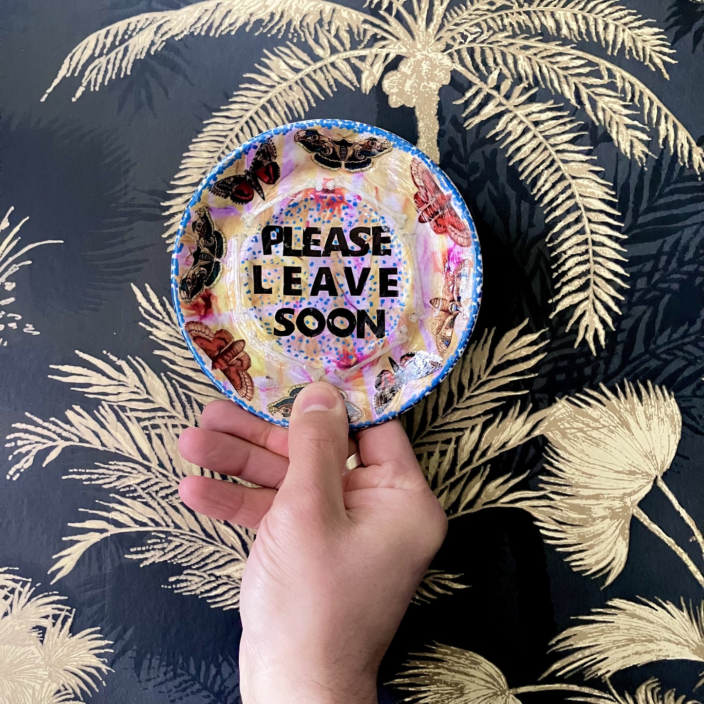 "Please Leave Soon" Trinket Dish by House of Frisson. Featuring some moths and the words "please leave soon", on yellow and pink background. Holding the dish against a wallpapered wall.