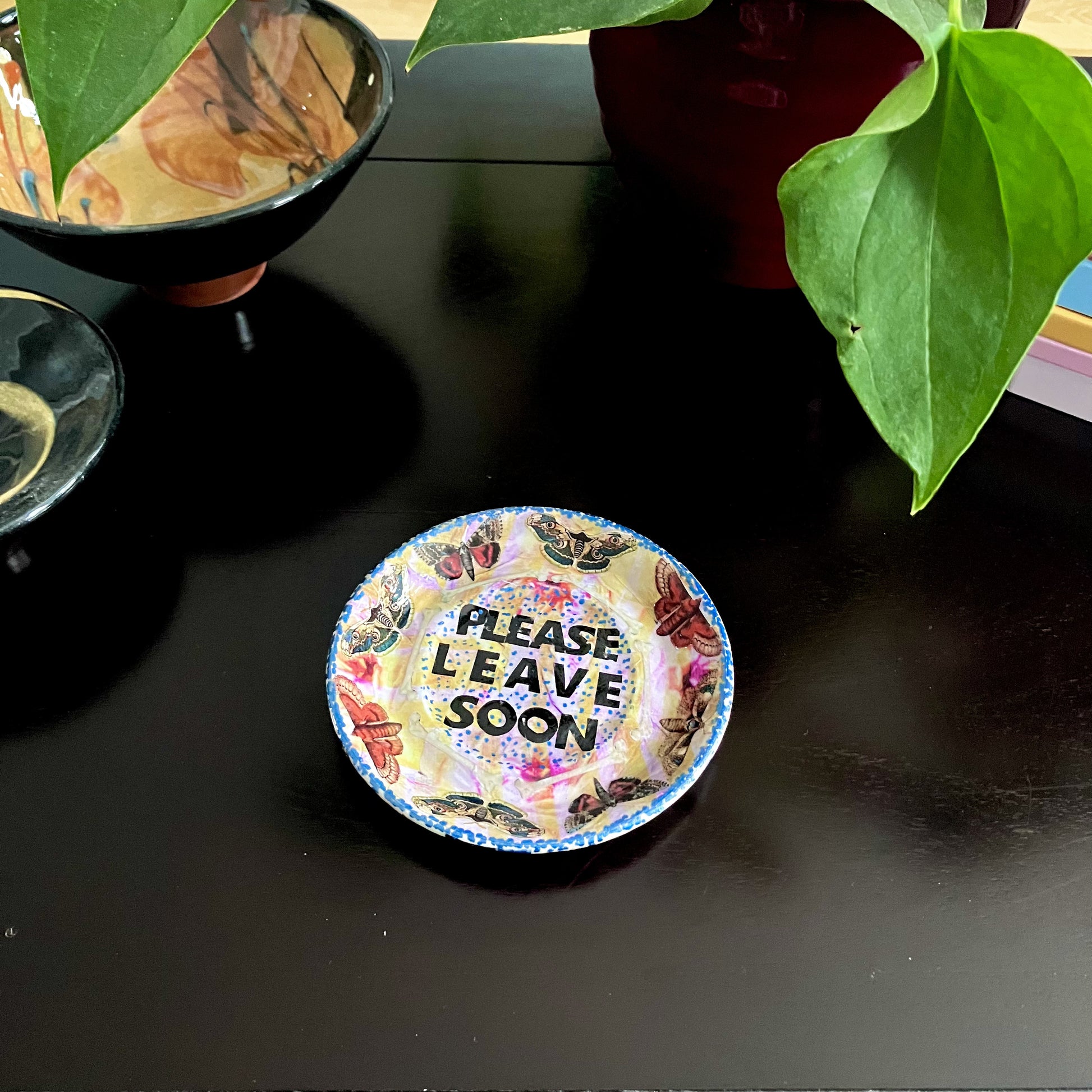 "Please Leave Soon" Trinket Dish by House of Frisson. Featuring some moths and the words "please leave soon", on yellow and pink background. Dish on a coffee table.
