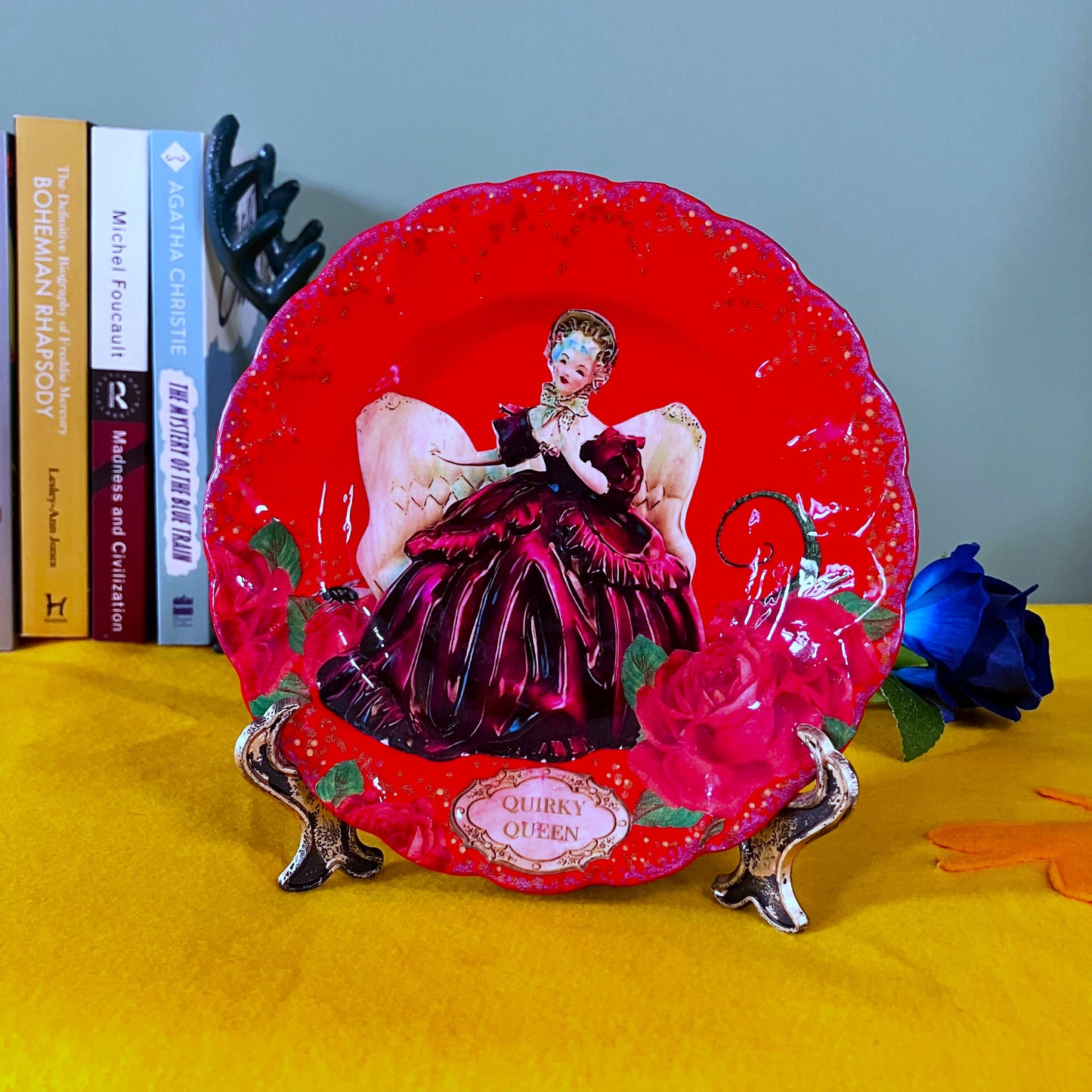 Quirky Queen Wall Plate by House of Frisson, featuring a kitsch porcelain figure of a lady surrounded by red roses and House of Frisson's creatures, against a red background. Plate on a plate stand resting on a table.