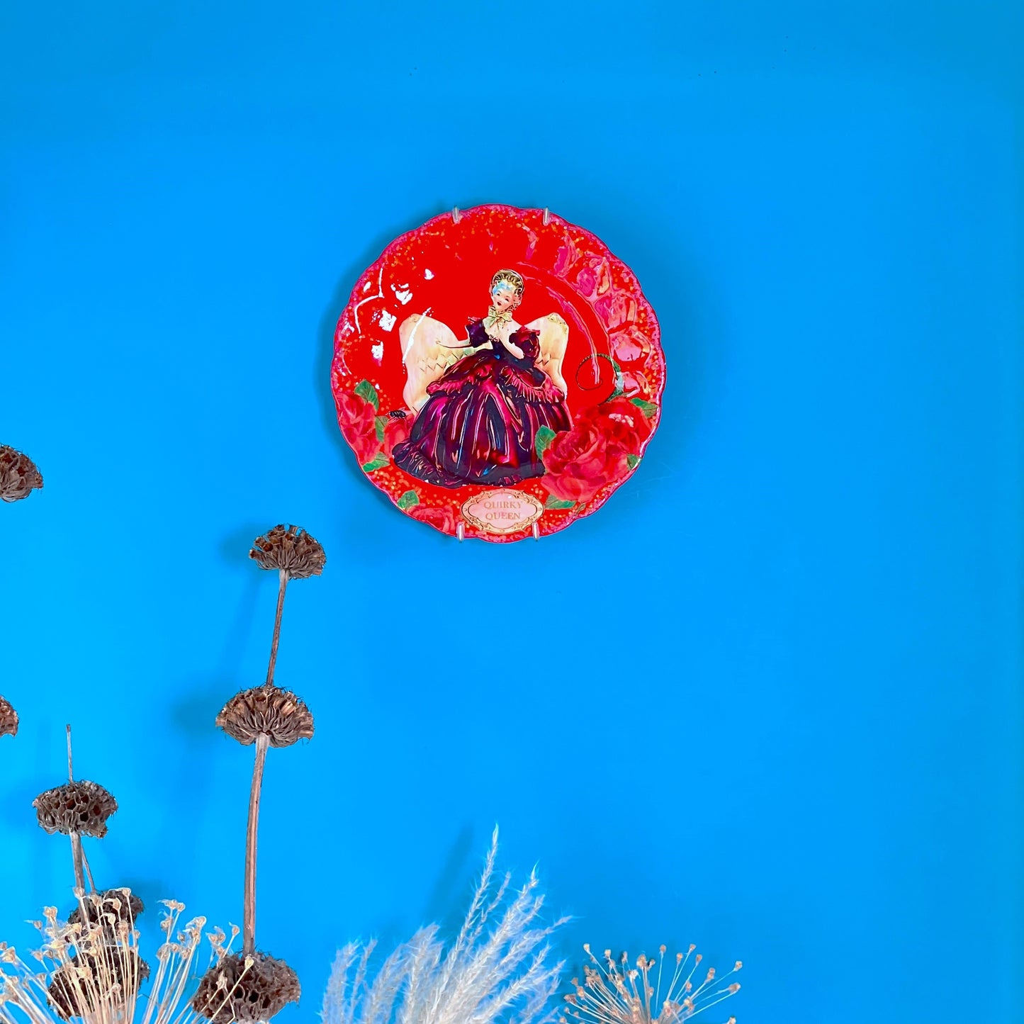 Quirky Queen Wall Plate by House of Frisson, featuring a kitsch porcelain figure of a lady surrounded by red roses and House of Frisson's creatures, against a red background. Plate hanging on a blue wall.
