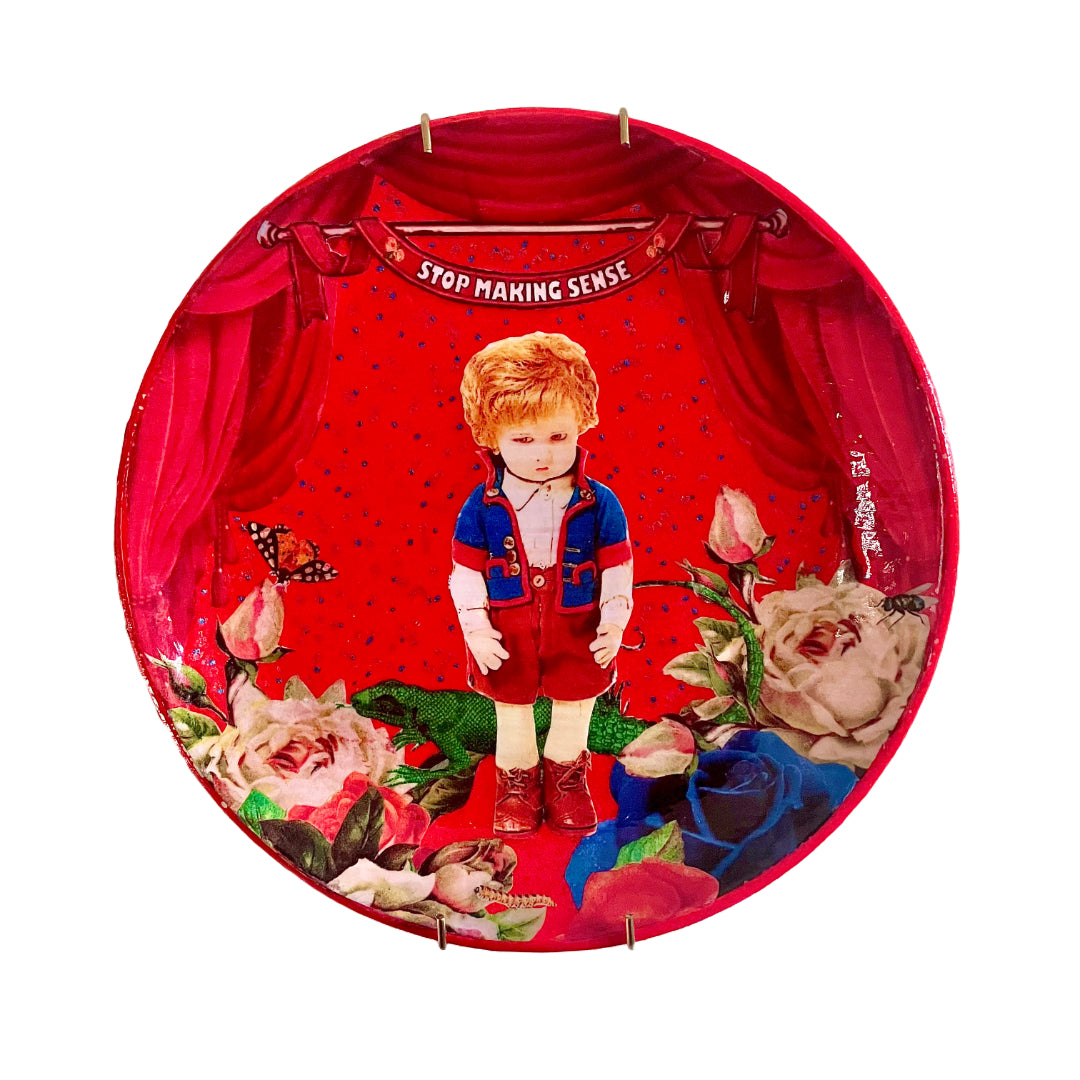 "Stop Making Sense" Wall Plate by House of Frisson, featuring a collage artwork of a felt male doll surrounded by roses, moths, and a lizard, framed by red curtains, on a red background.