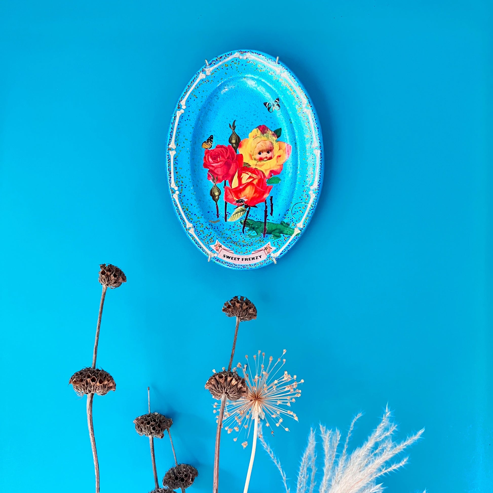 "Sweet Frenzy" Upcycled Wall Plate by House of Frisson, featuring some vintage flowers, moths, a fly, and a lizard, against a turquoise background and a frame of bones. Plate hanging on a turquoise wall.