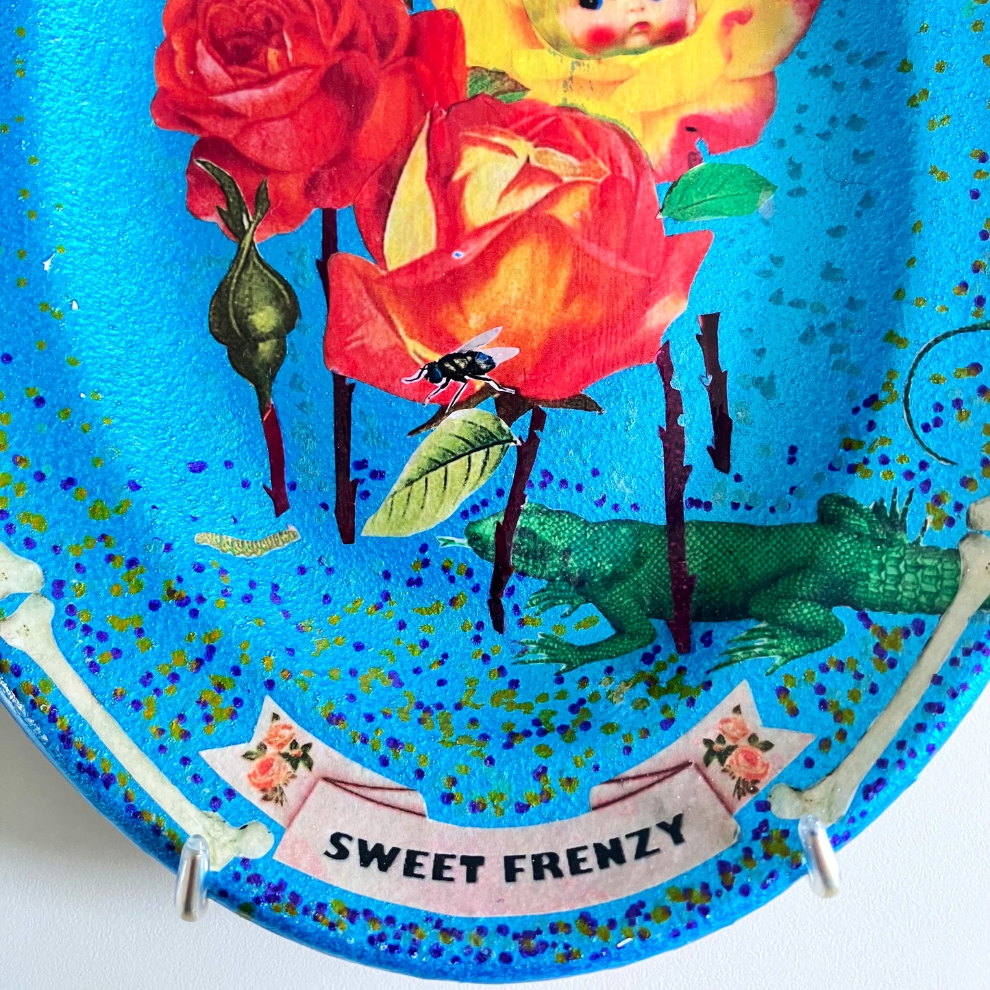 "Sweet Frenzy" Upcycled Wall Plate by House of Frisson, featuring some vintage flowers, moths, a fly, and a lizard, against a turquoise background and a frame of bones. Closeup detail showing the collage.