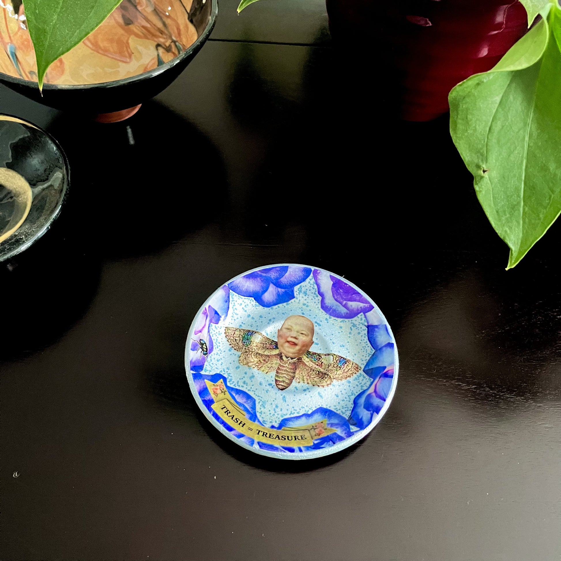 "Trash = Treasure" Trinket Dish by House of Frisson. Featuring a collage of a moth with a doll head, framed with blue roses. Showing dish on a coffee table.