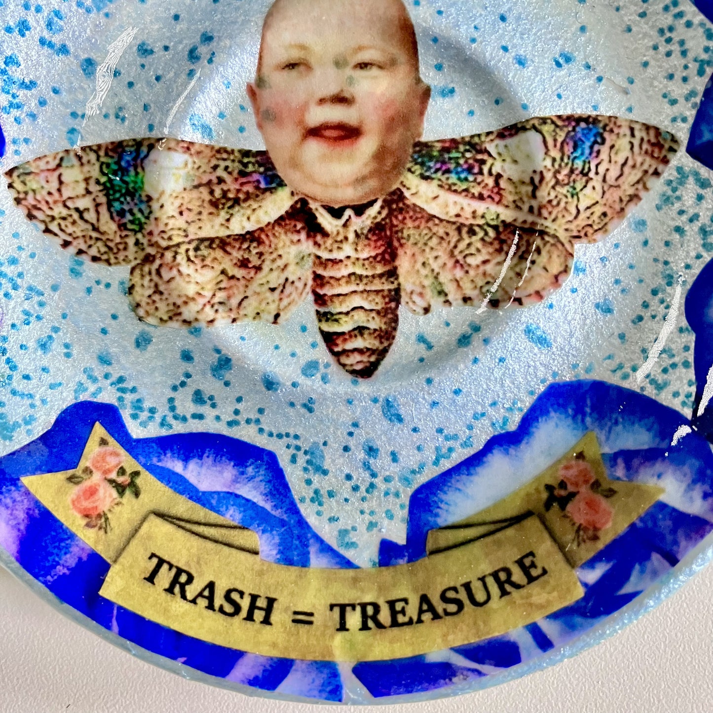 "Trash = Treasure" Trinket Dish by House of Frisson. Featuring a collage of a moth with a doll head, framed with blue roses. Closeup detail.