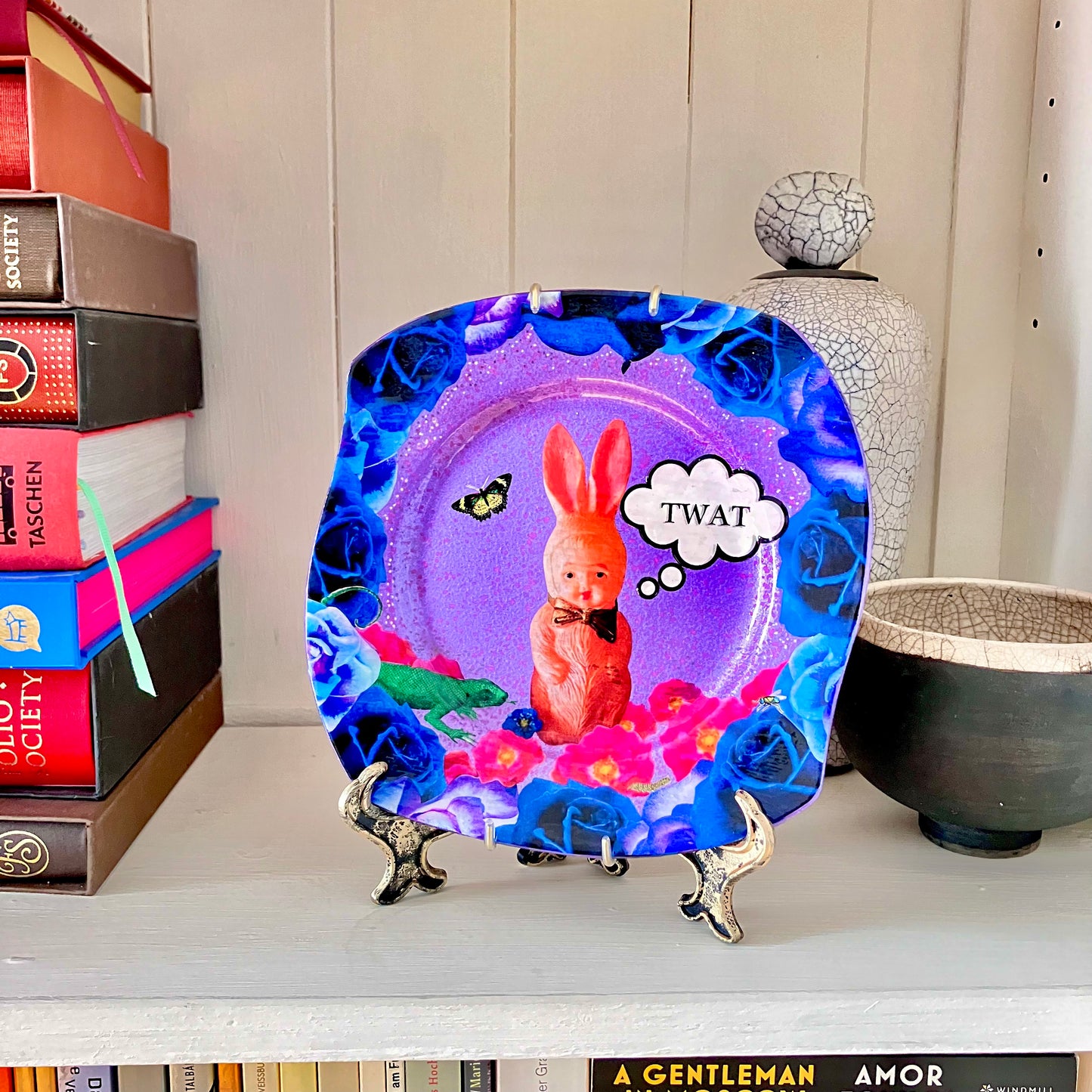 "Twat" Wall Plate by House of Frisson on a plate stand resting on a bookshelf.