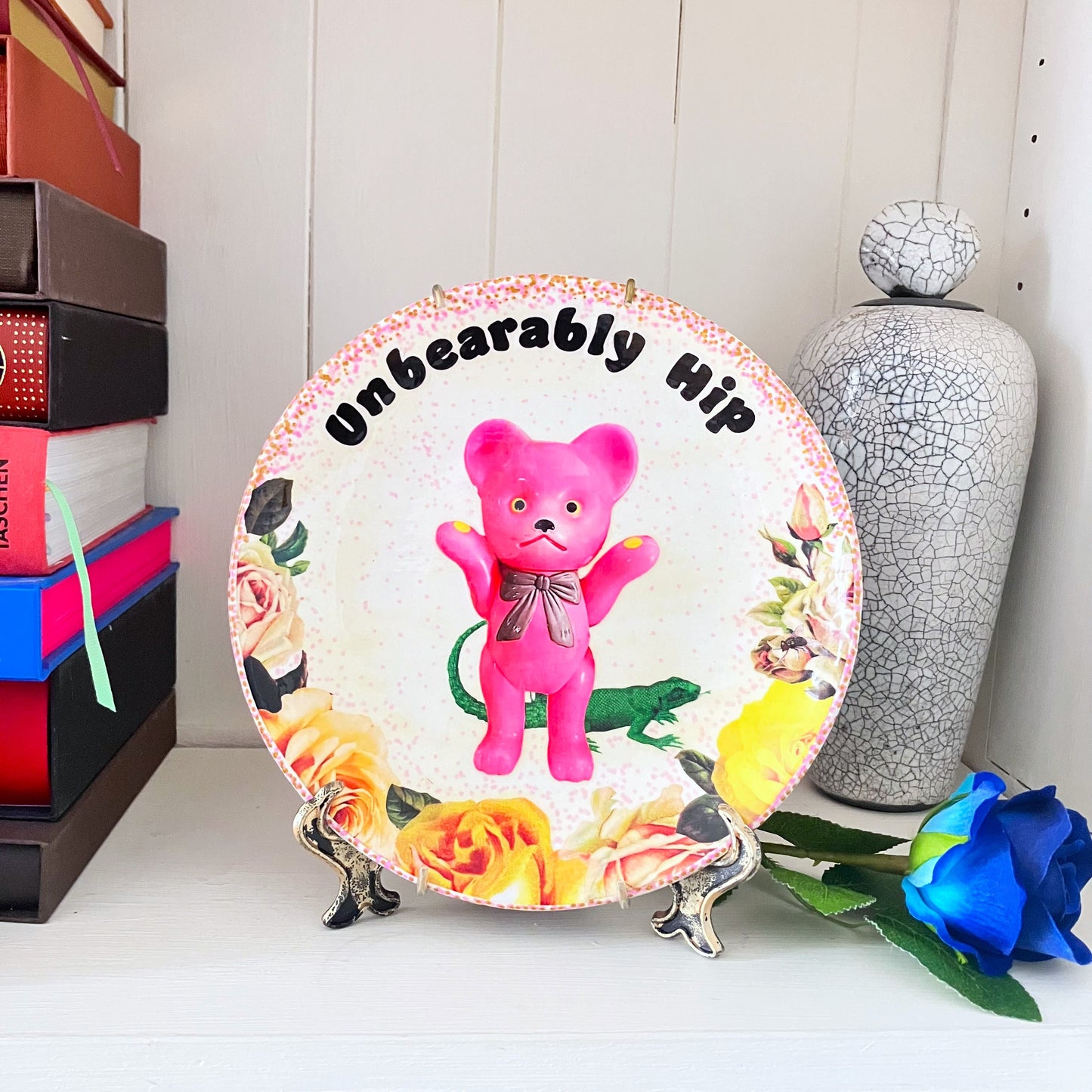 White Upcycled Wall Plate featuring a kitschy pink bear surrounded by whimsical creatures and roses, making it "Unbearably Hip." Plate on a stand resting on a shelf.