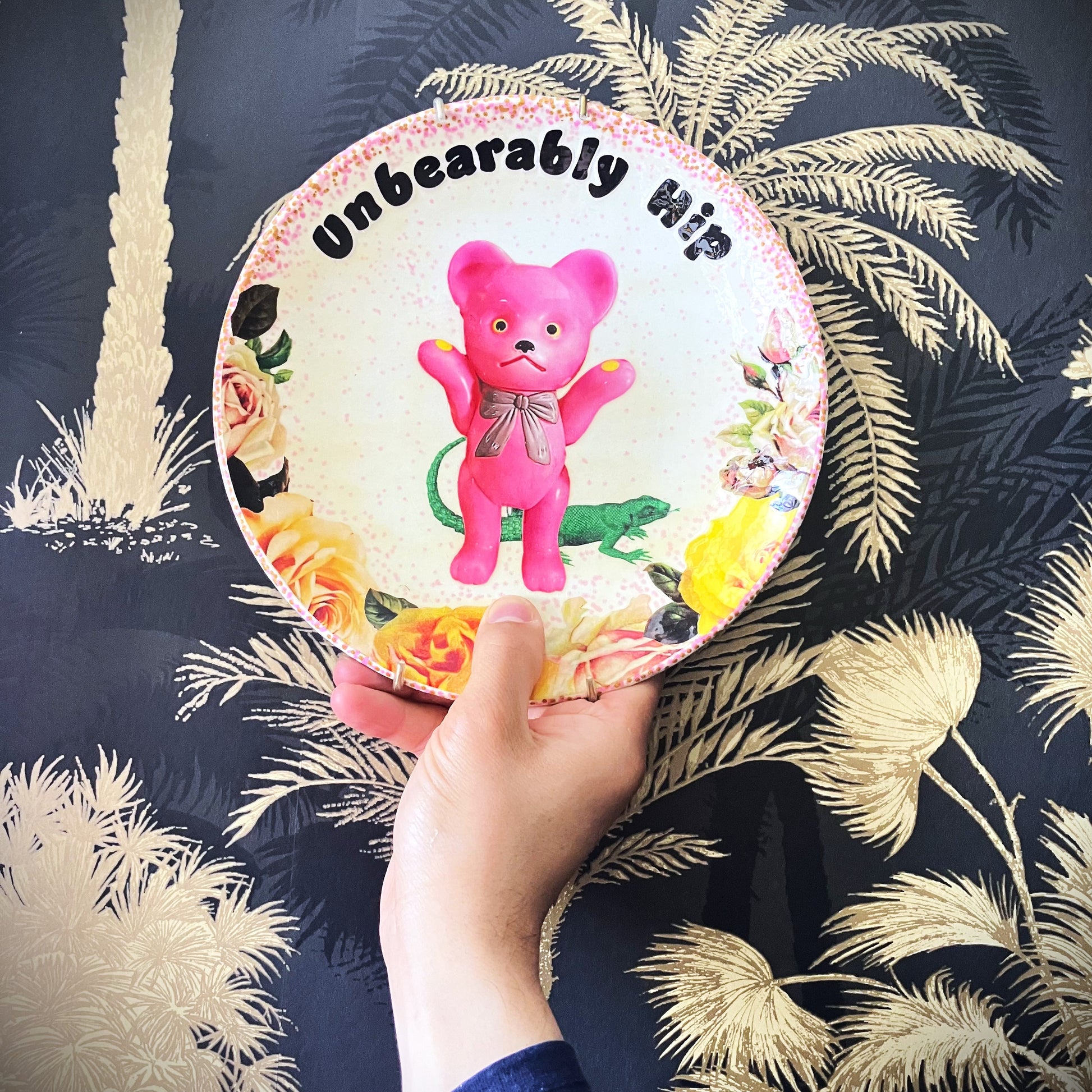 White Upcycled Wall Plate featuring a kitschy pink bear surrounded by whimsical creatures and roses, making it "Unbearably Hip." Plate on hand.