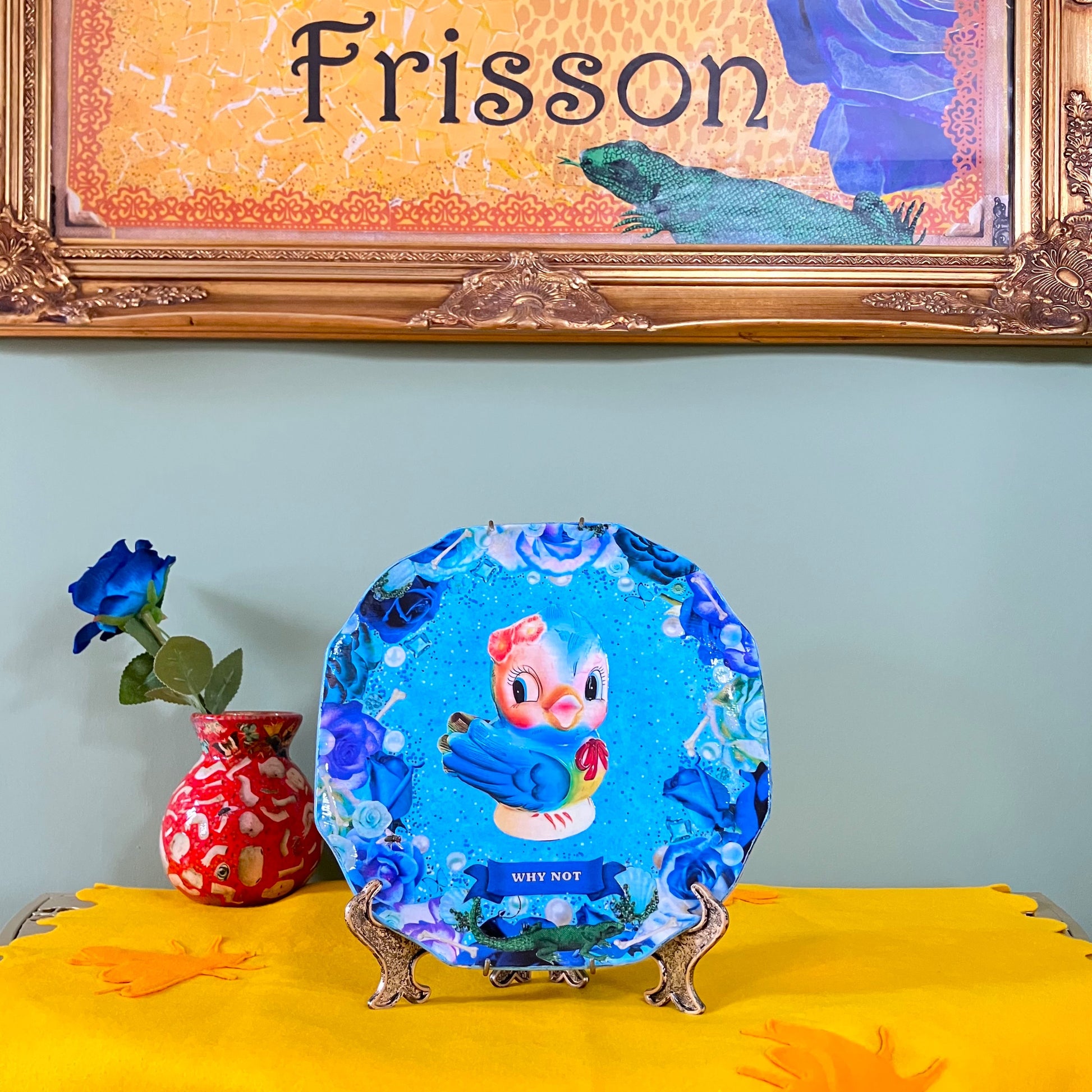 Blue upcycled wall plate by House of Frisson featuring a collage of a vintage ceramic bird image with blue roses, pearls, and bones. Plate on a plate stand resting on a table.