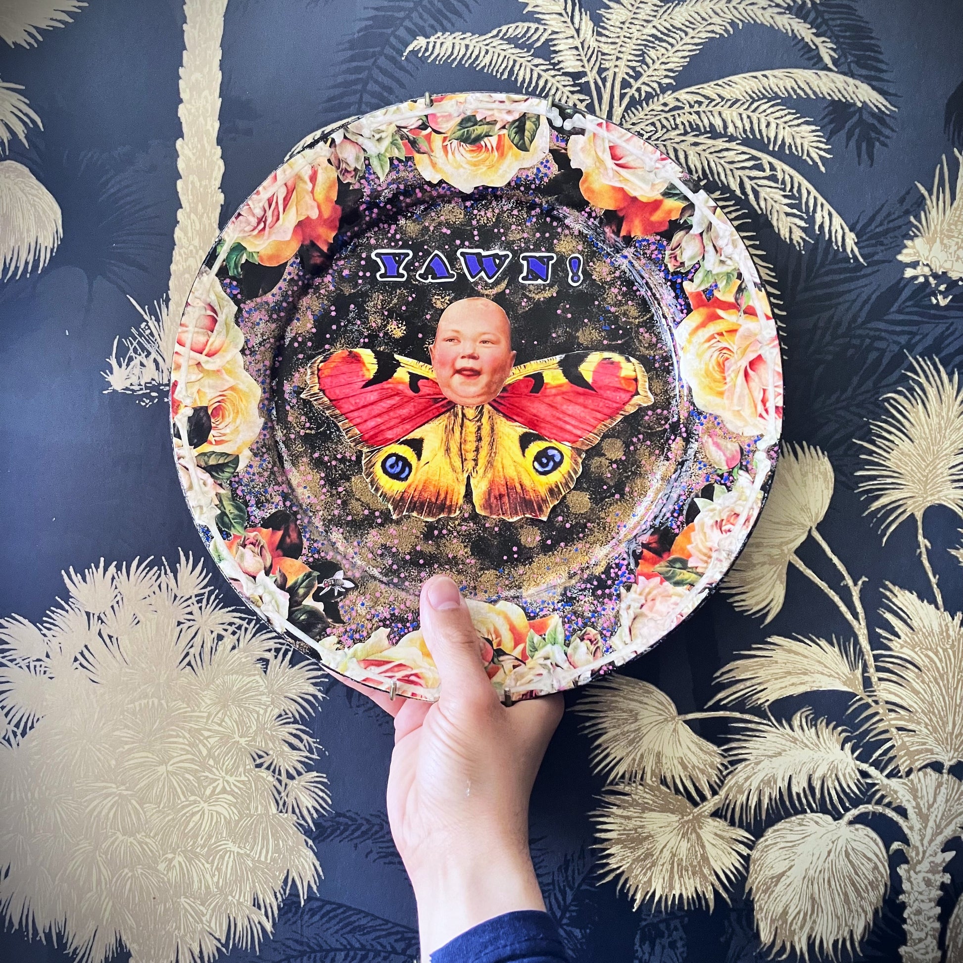 Upcycled Wall Plate by House of Frisson. Featuring a unique collage of a moth with a doll face, surrounded by delicate roses and bones, this quirky piece is anything but ordinary. Holding the plate.