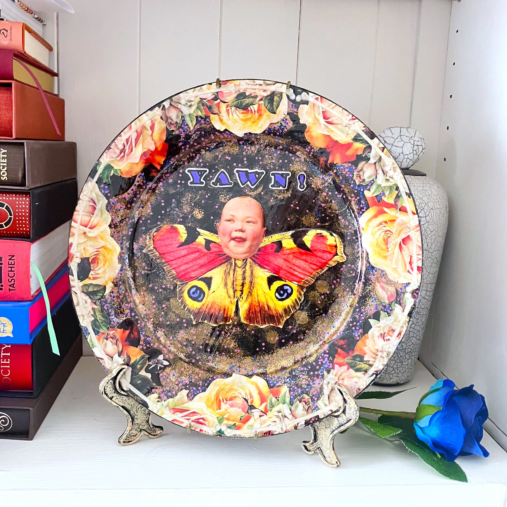 Upcycled Wall Plate by House of Frisson. Featuring a unique collage of a moth with a doll face, surrounded by delicate roses and bones, this quirky piece is anything but ordinary. Plate on plate holder resting on a shelf.