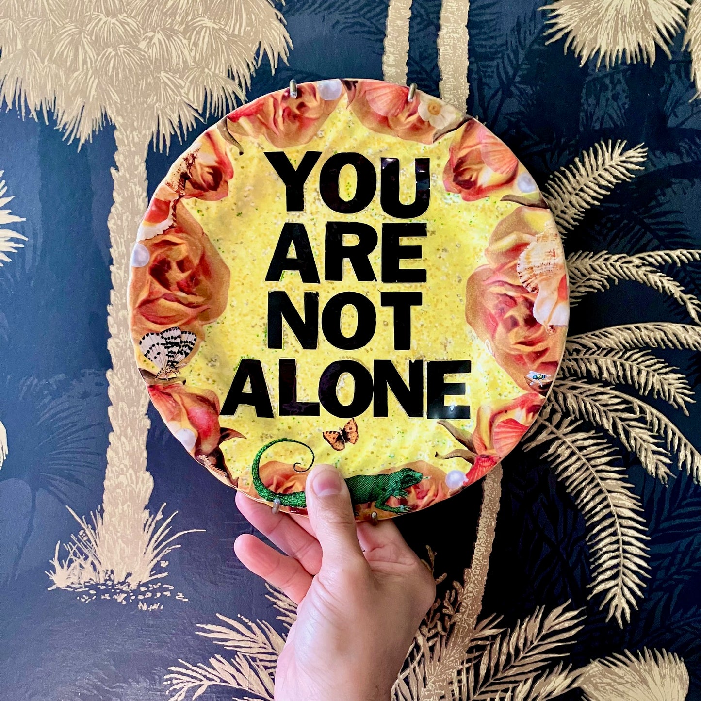 "You Are Not Alone" one-of-a-kind collage artwork Wall Plate by House of Frisson. Featuring the words "You Are Not Alone" framed by roses, pearls, and moths, on a yellow background. Holding the plate against a wallpaper.