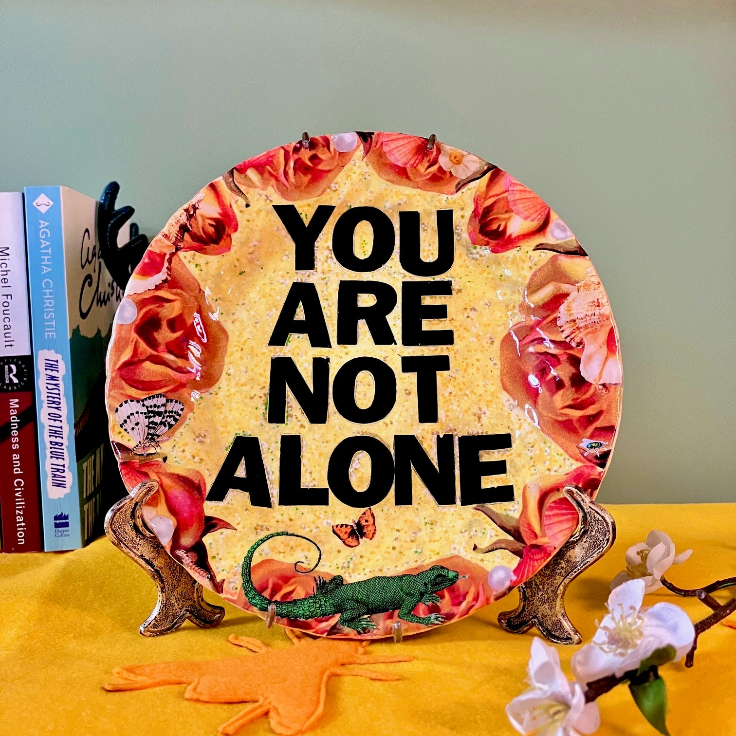 "You Are Not Alone" one-of-a-kind collage artwork Wall Plate by House of Frisson. Featuring the words "You Are Not Alone" framed by roses, pearls, and moths, on a yellow background. Plate on a plate stand resting on a table.