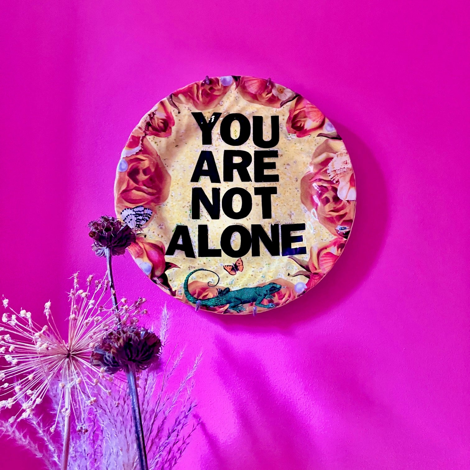 "You Are Not Alone" one-of-a-kind collage artwork Wall Plate by House of Frisson. Featuring the words "You Are Not Alone" framed by roses, pearls, and moths, on a yellow background. Plate hanging on a pink wall.