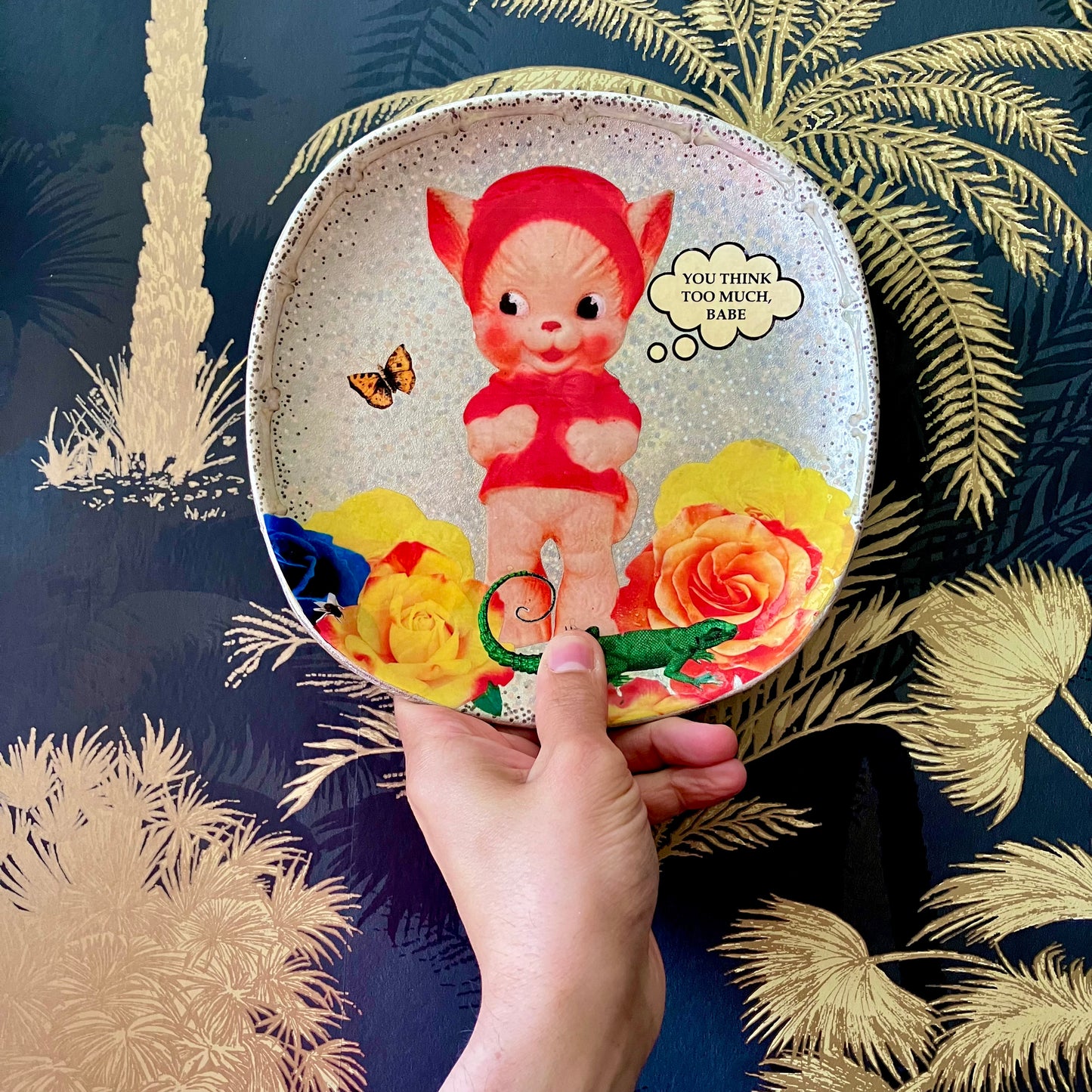 "You Think Too Much, Babe" Silver upcycled wall plate by House of Frisson, featuring a kitsch squeaky toy, some colourful roses and House of Frisson's creatures. Plate on hand.