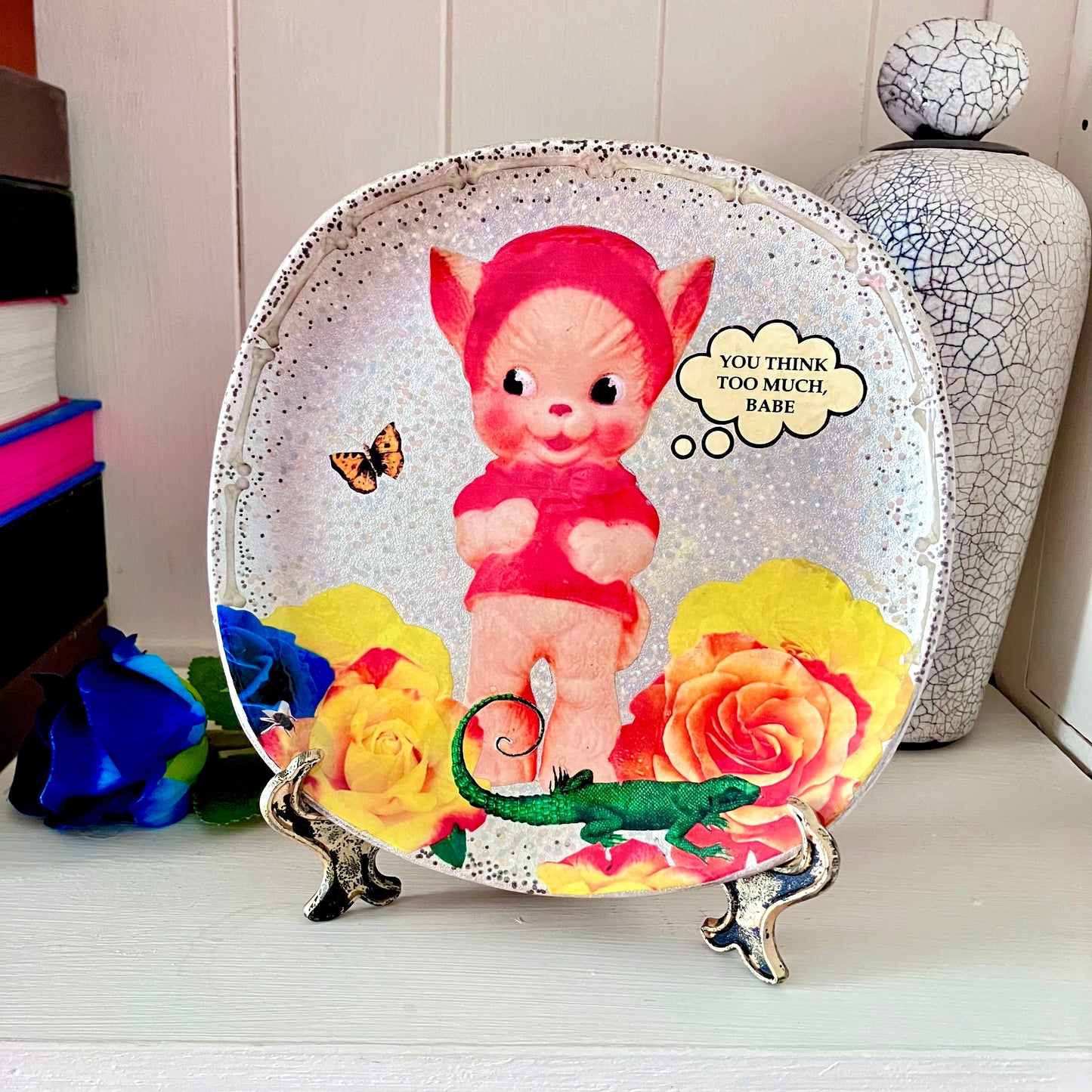 "You Think Too Much, Babe" Silver upcycled wall plate by House of Frisson, featuring a kitsch squeaky toy, some colourful roses and House of Frisson's creatures. Plate on a plate stand resting on a shelf.