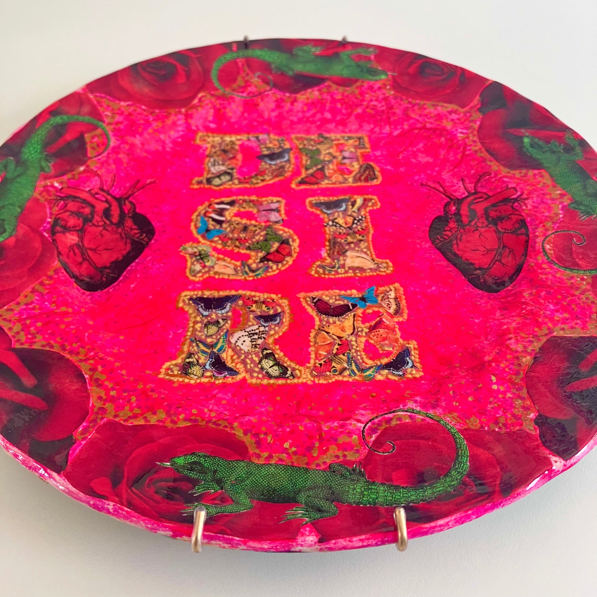 "Desire" Wall Plate by House of Frisson, featuring a collage with the word "desire" written with moths, and a frame of red roses and green lizards, on a hot pink background. Closeup details.